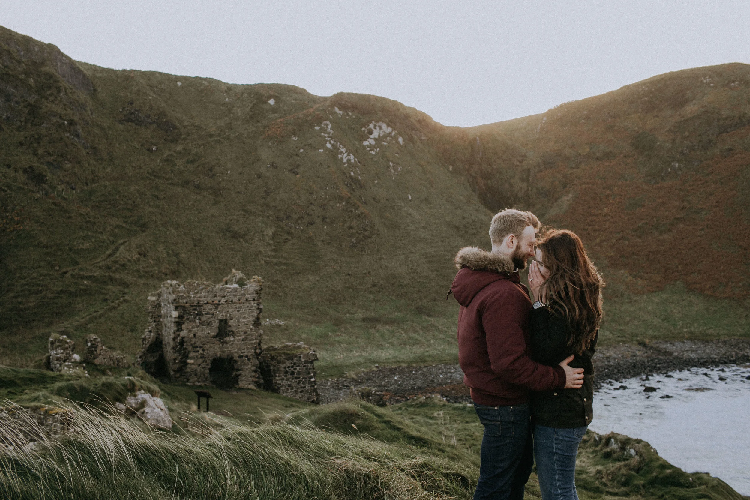 A couple standing close together on a grassy hillside near a body of water, with old stone ruins and green, rolling hills in the background.