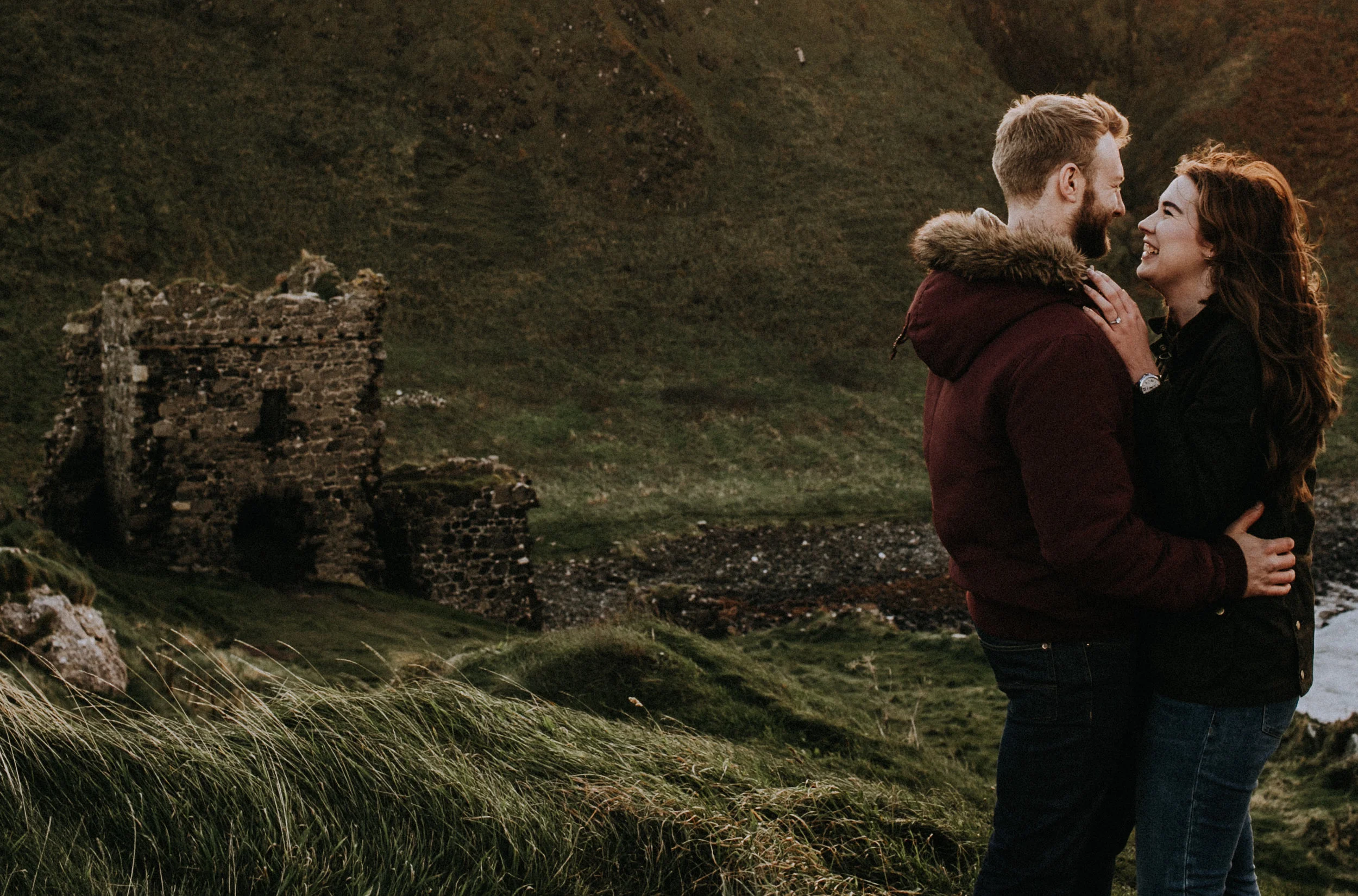 A couple embraces and looks at each other in a green hilly landscape with an old stone ruins in the background during sunset.