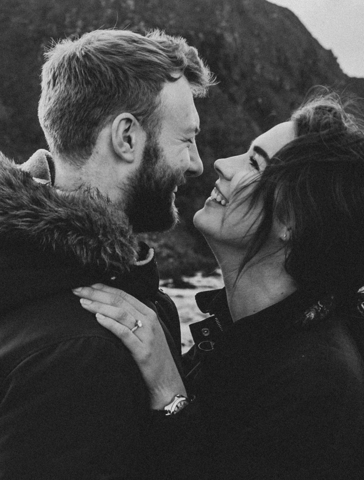 A black-and-white photo of a smiling couple looking at each other, close together outdoors with a mountain and cloudy sky in the background.