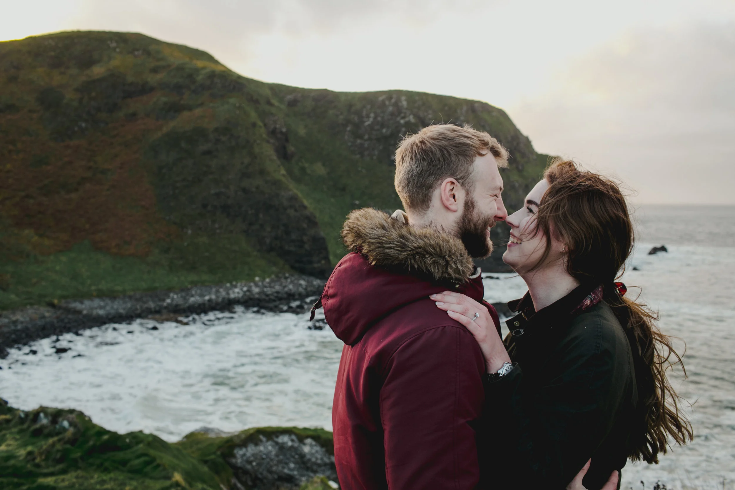 A couple is standing on a rocky coastline with the ocean and green cliffs in the background. They are smiling and very close, about to kiss.