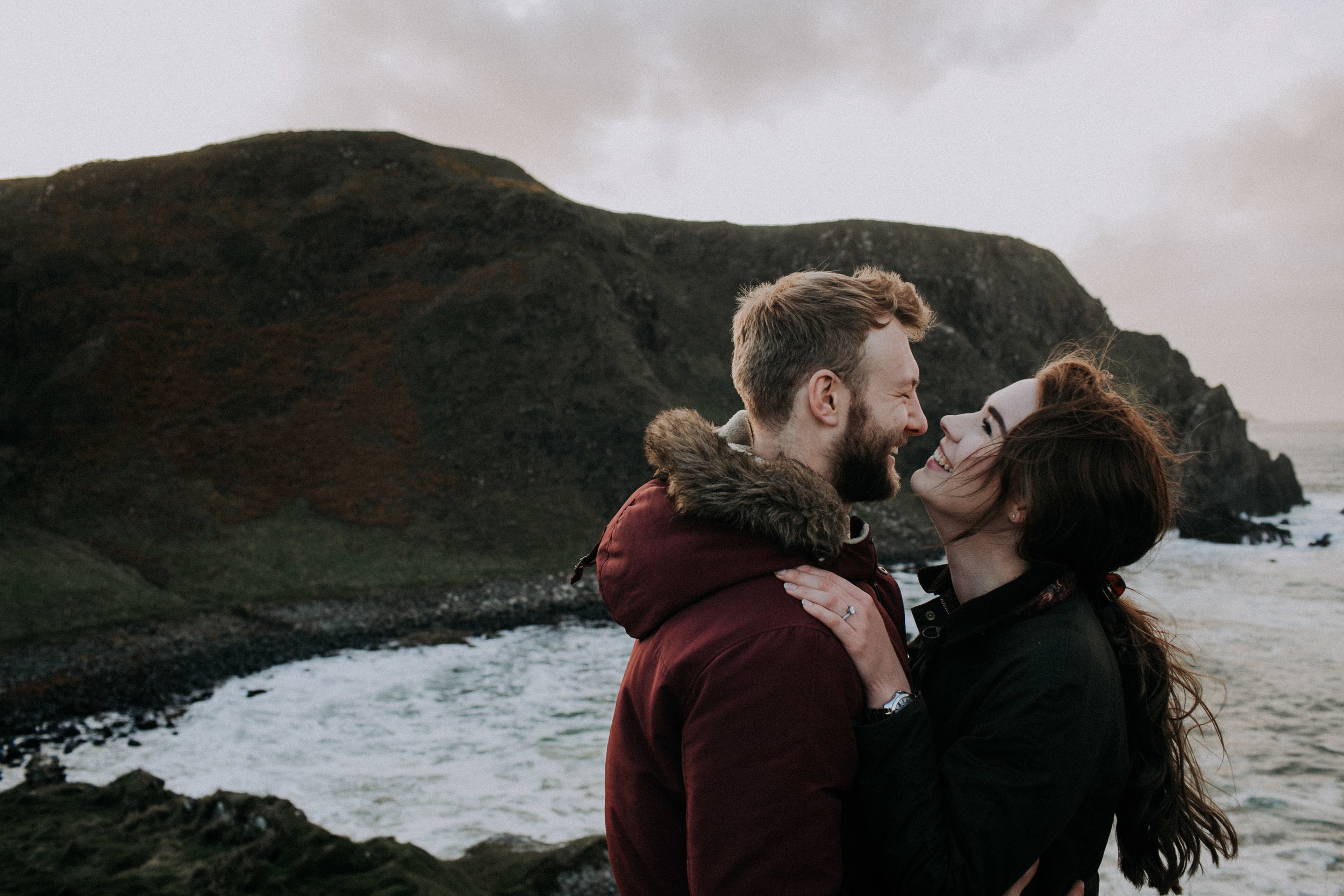 A happy couple is embracing and smiling at each other near the coastline with dark cliffs and the ocean in the background.