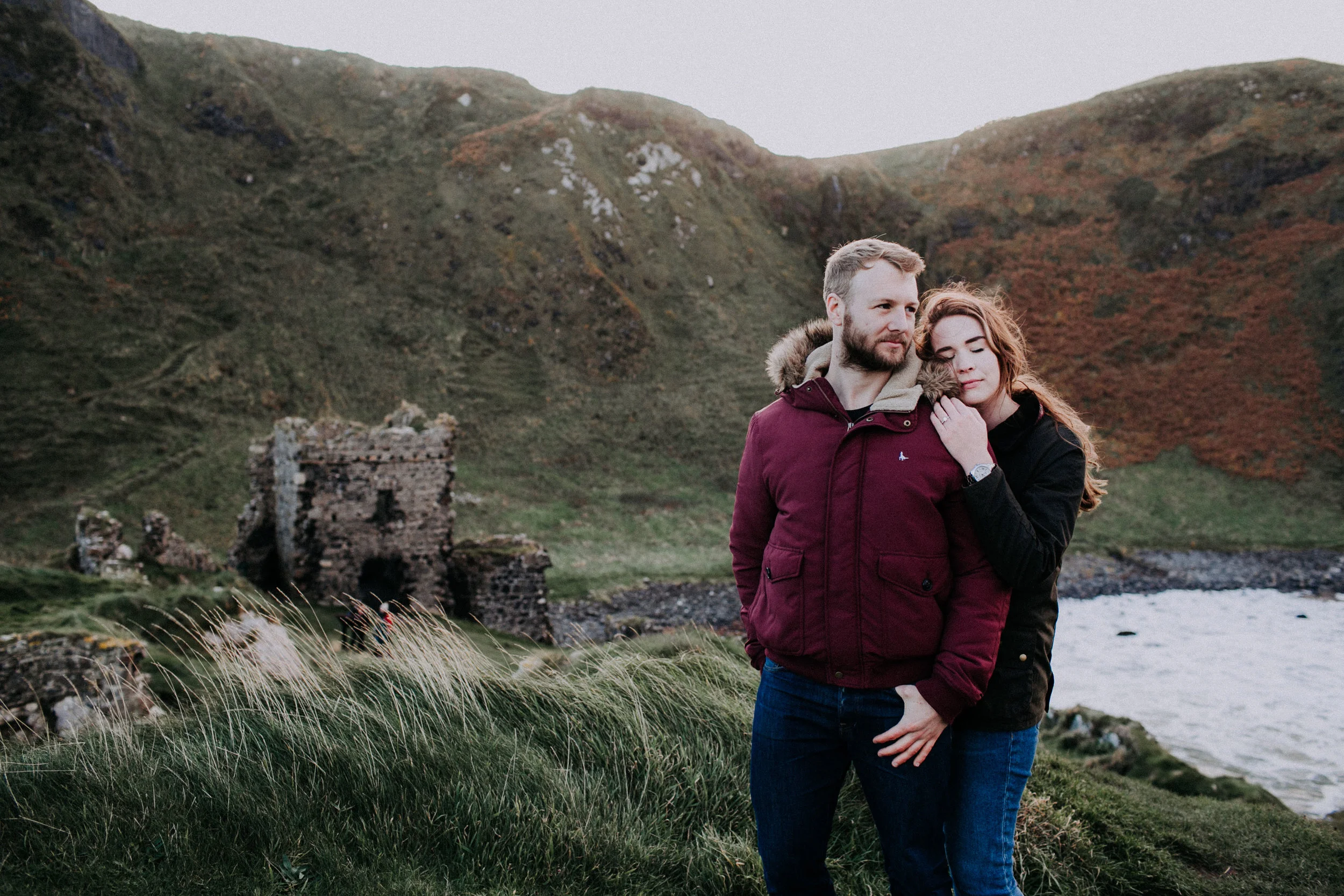 A couple standing on grassy terrain near a body of water, with ancient ruins and hills in the background.