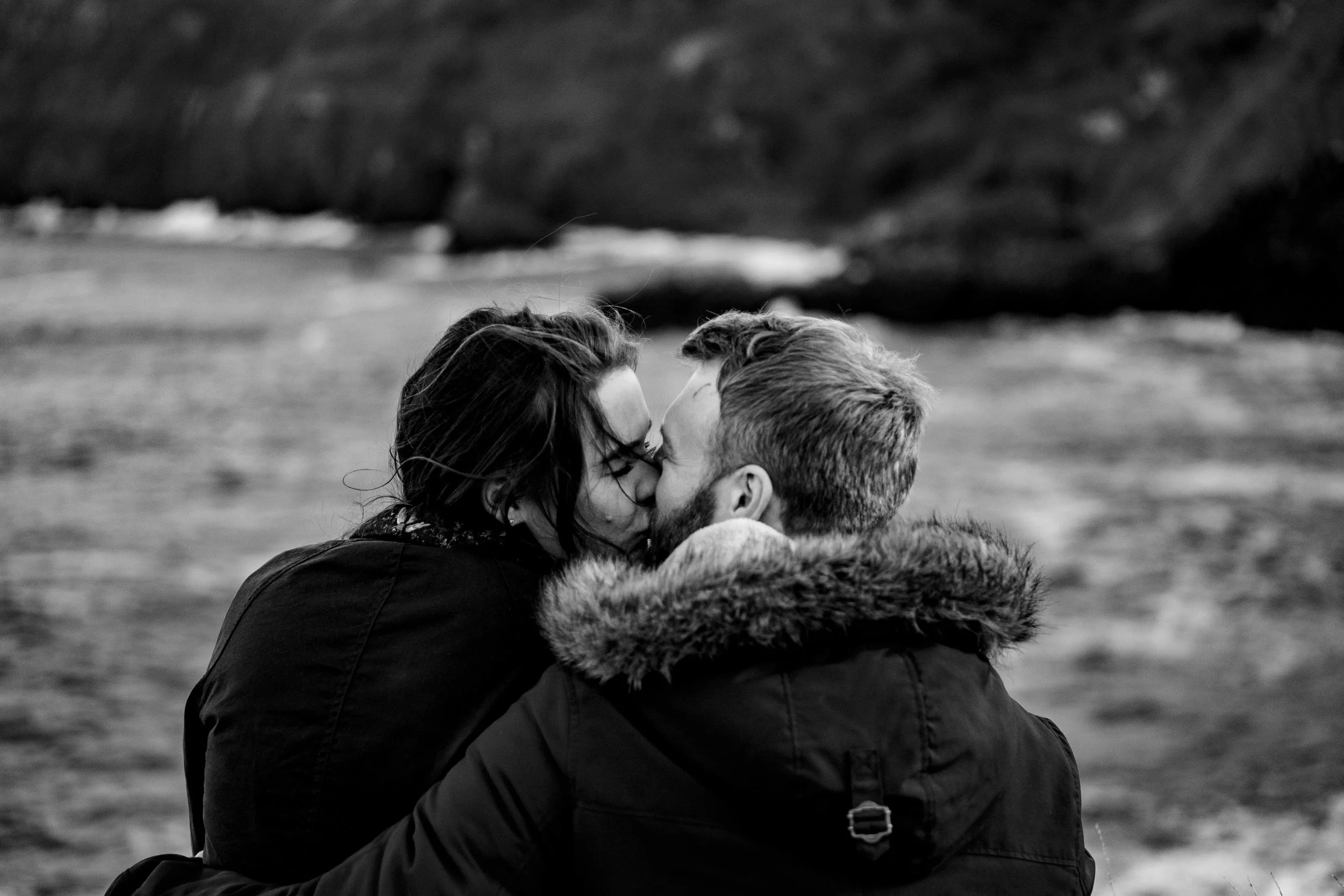 A black and white photo of a couple kissing outdoors near the water, with a rocky shoreline or mountain in the background.