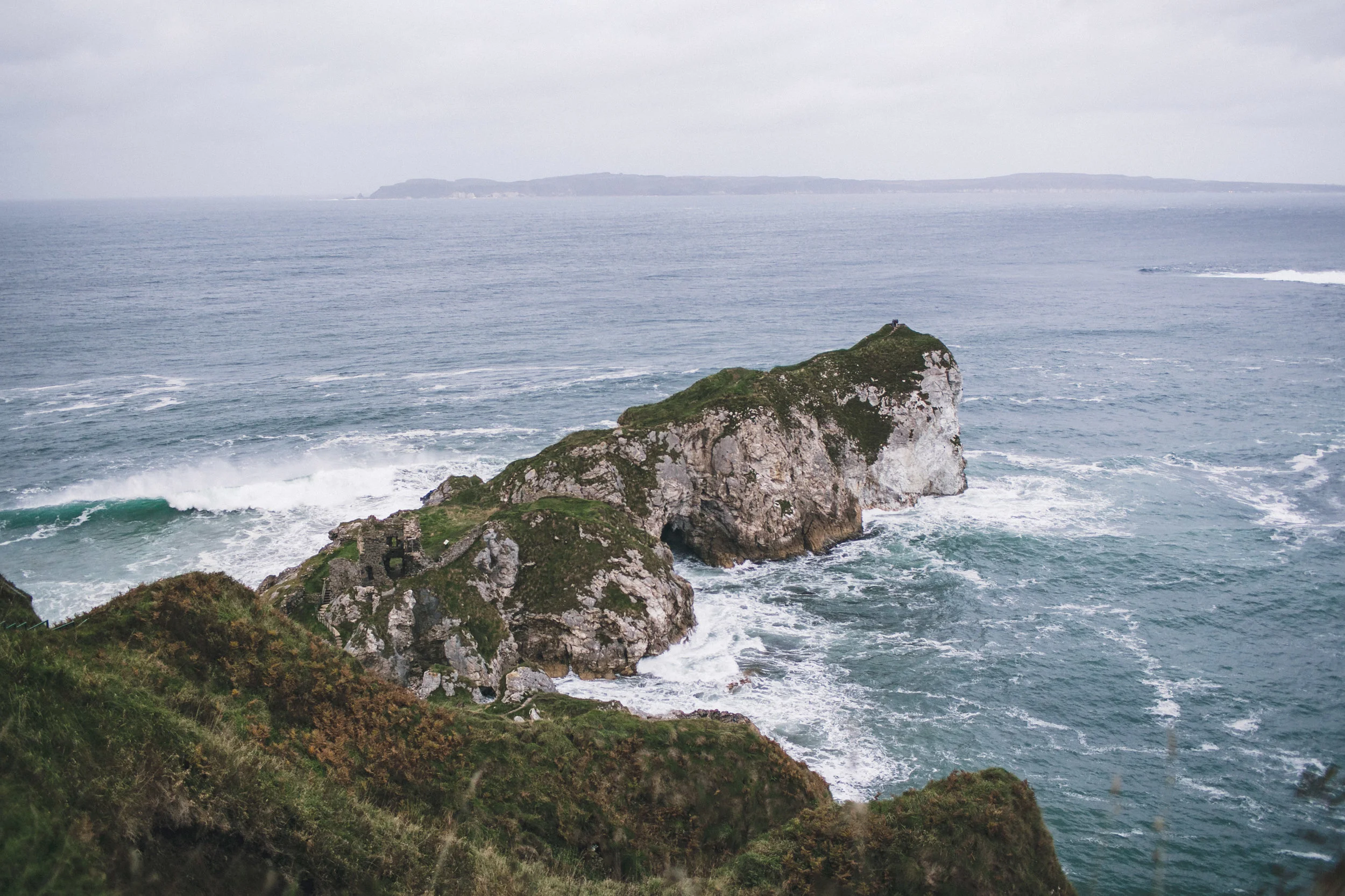 A rocky island with green vegetation surrounded by ocean waves, with a distant landmass on the horizon under overcast sky.