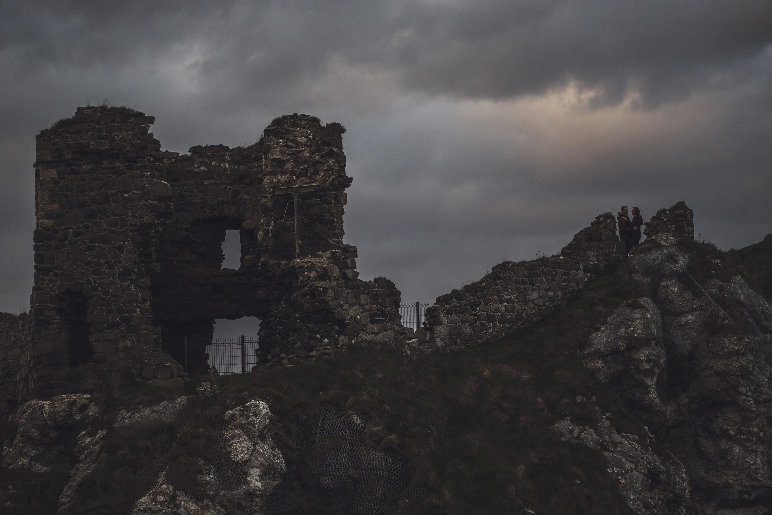 Ruins of an ancient stone castle on a rocky hill under a dark, cloudy sky, with two people standing on the hilltop.