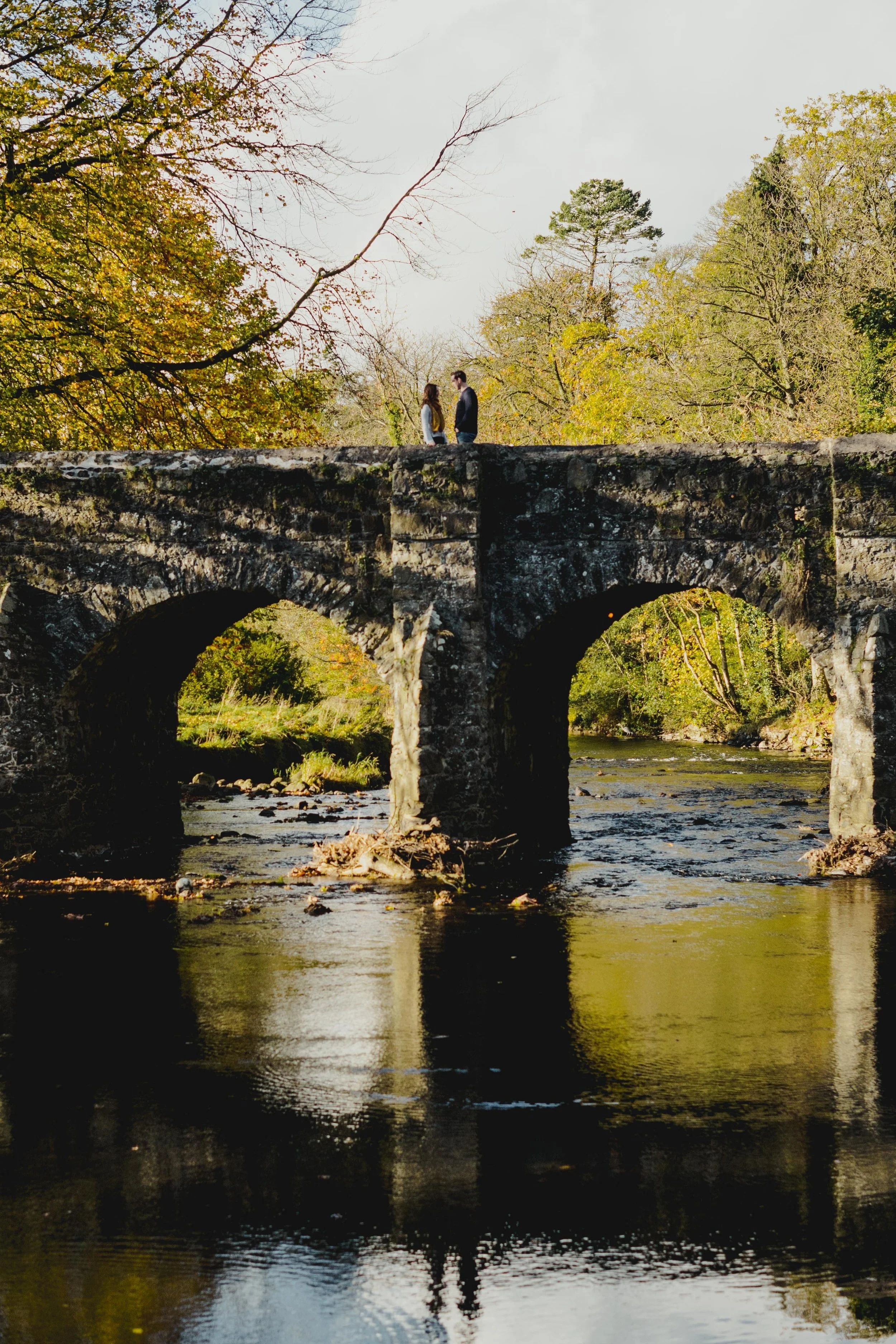 A couple standing on an old stone bridge over a river, surrounded by trees in fall colors.