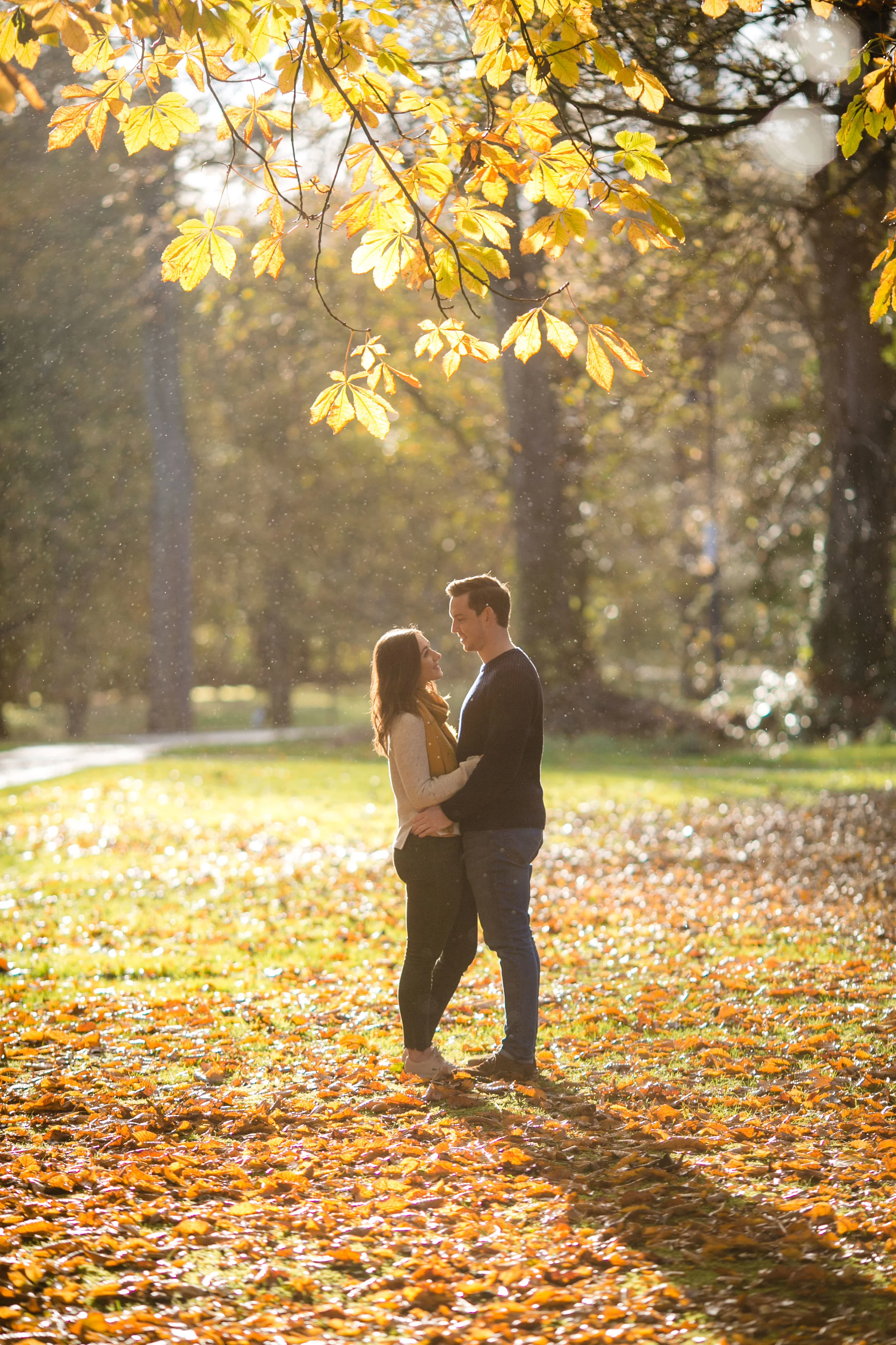 A young couple stands closely together in a park on a fall day, surrounded by fallen leaves and golden sunlight filtering through trees.