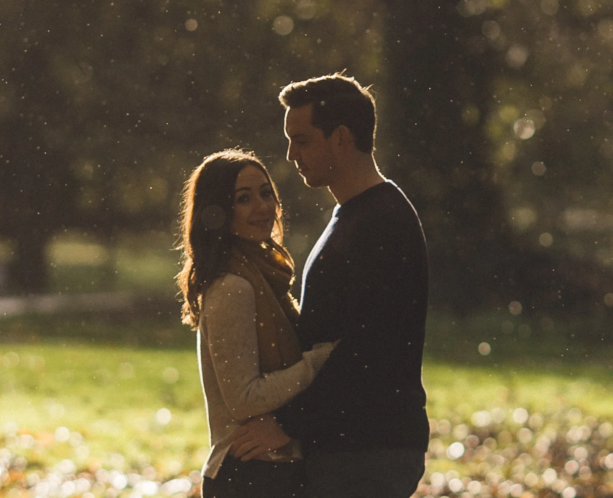 A couple standing close together outdoors at sunset, with the woman smiling at the camera and the man looking down, surrounded by glowing particles and bokeh lights.