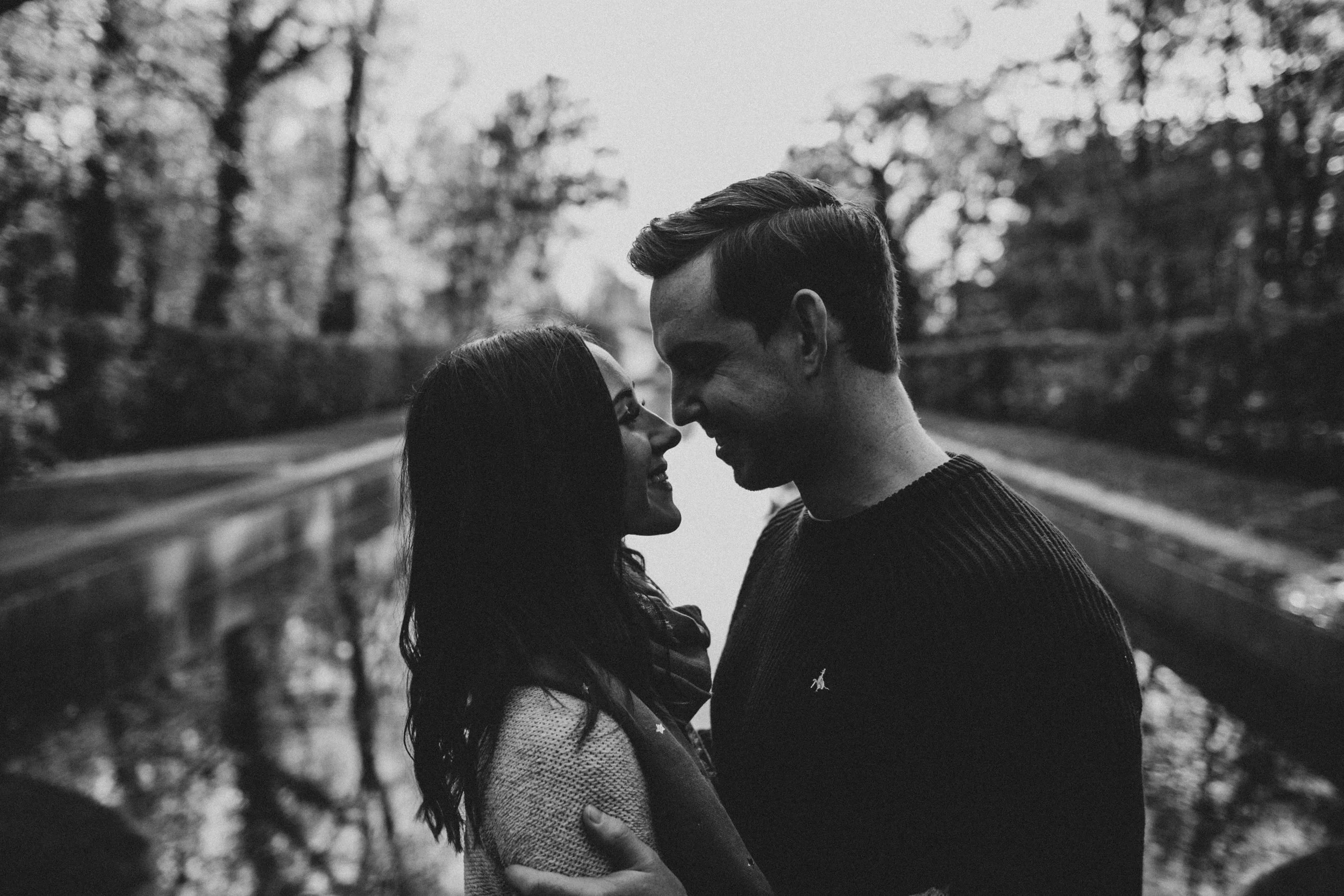 A black and white photo of a couple standing close together outdoors, faces nearly touching, in a romantic moment surrounded by trees.