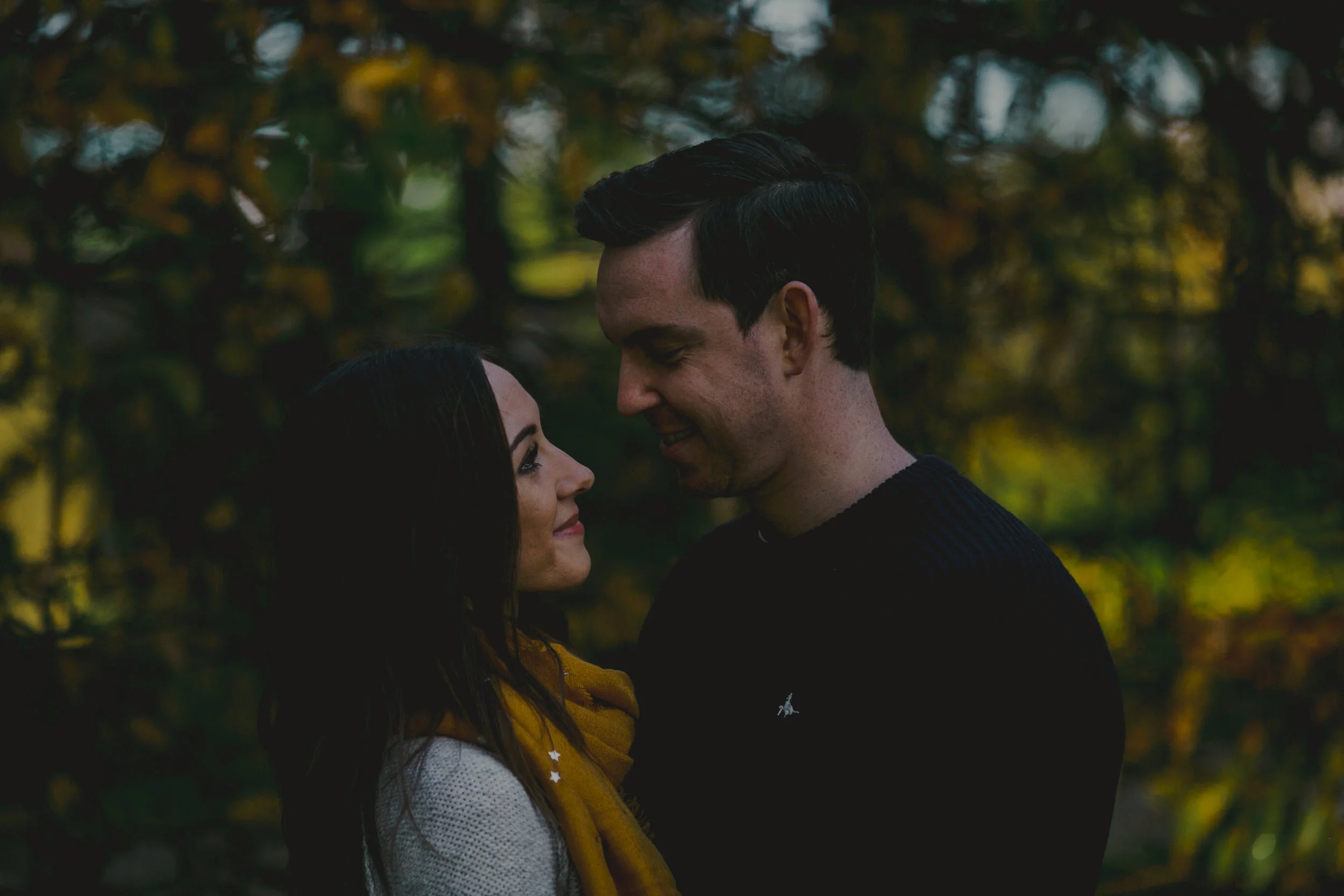 A couple standing close together outdoors with autumn trees in the background, smiling and gazing at each other.