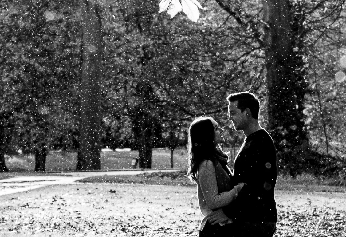 A black and white photo of a couple standing close and smiling at each other outdoors, with snow falling around them. Trees are in the background.