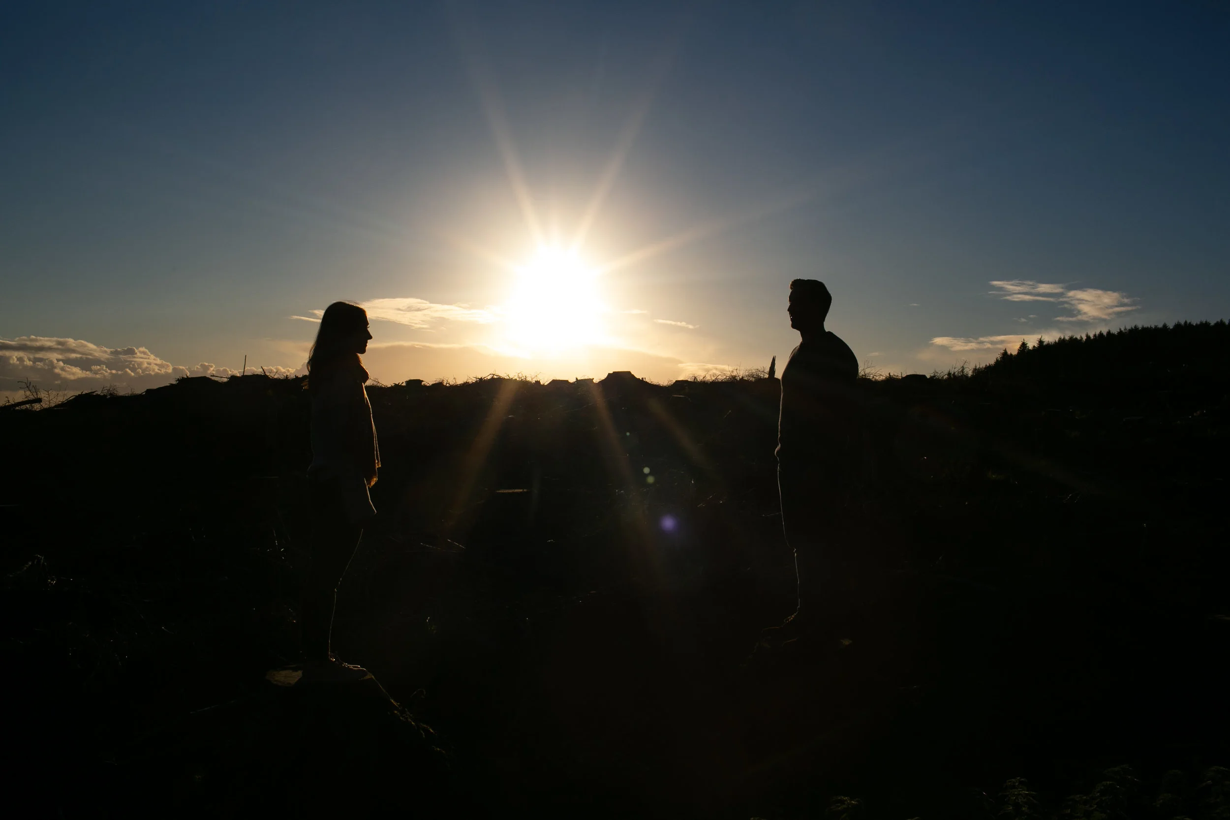 Silhouettes of a man and a woman facing each other during sunset with the sun low in the sky, casting a glow over the landscape.