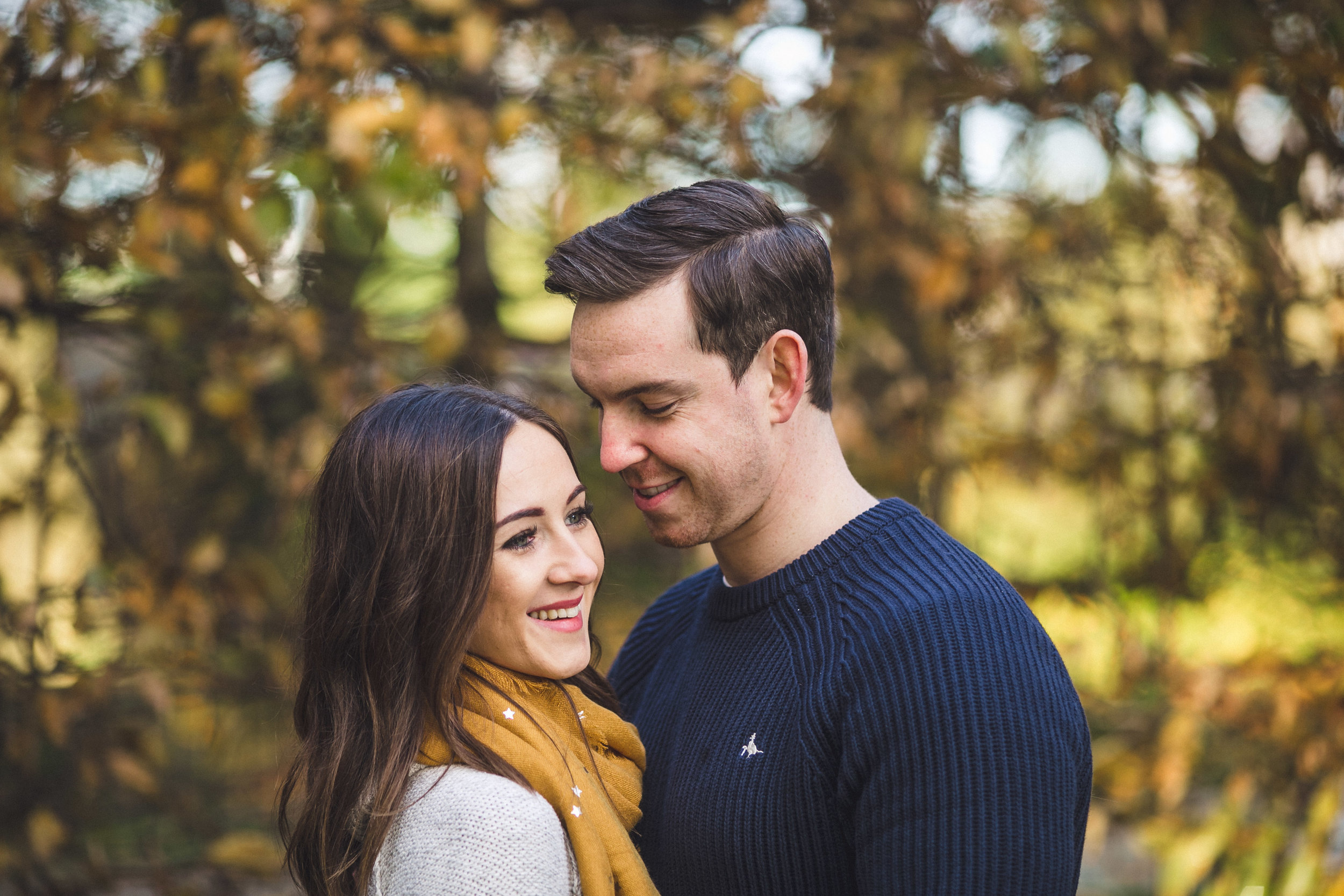 A young couple standing close together outdoors during autumn, surrounded by trees with fall foliage. The woman has long brown hair and is wearing a white sweater and a mustard yellow scarf. The man has short dark hair and is wearing a dark blue swea
