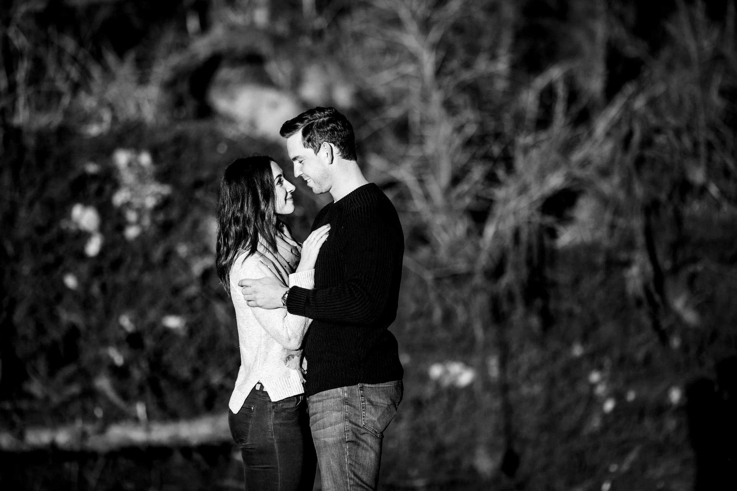 A black-and-white photo of a young couple standing close together, smiling and looking into each other's eyes outdoors at night.