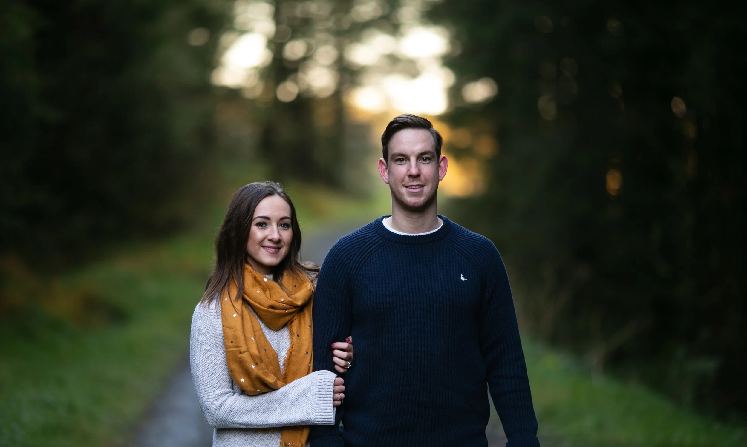 A young couple standing arm-in-arm outdoors on a forested trail during sunset, smiling at the camera. Ballyboley Forest