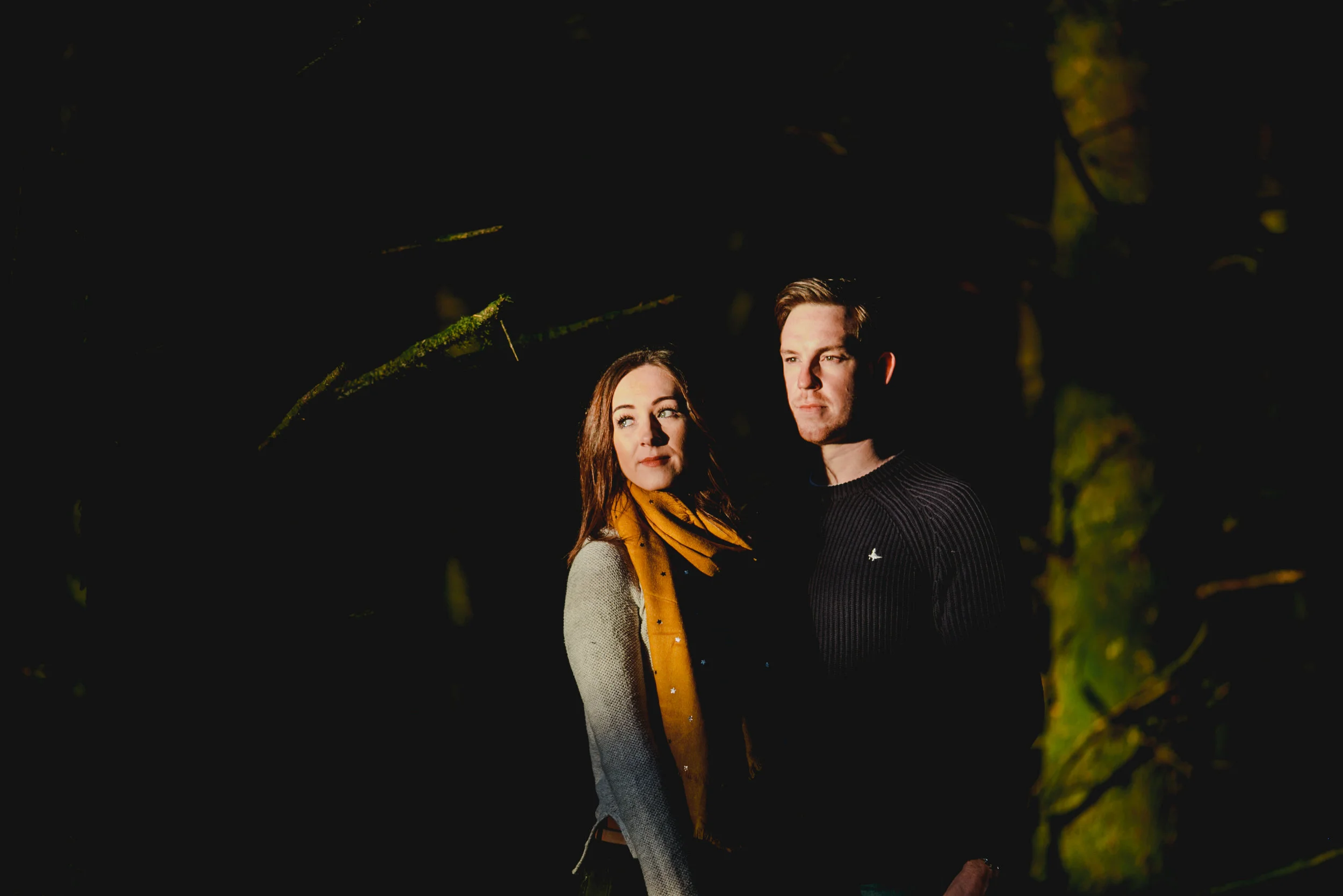 A man and a woman standing outdoors at night, illuminated by a light source, with dark background and tree branches. Couples Session
