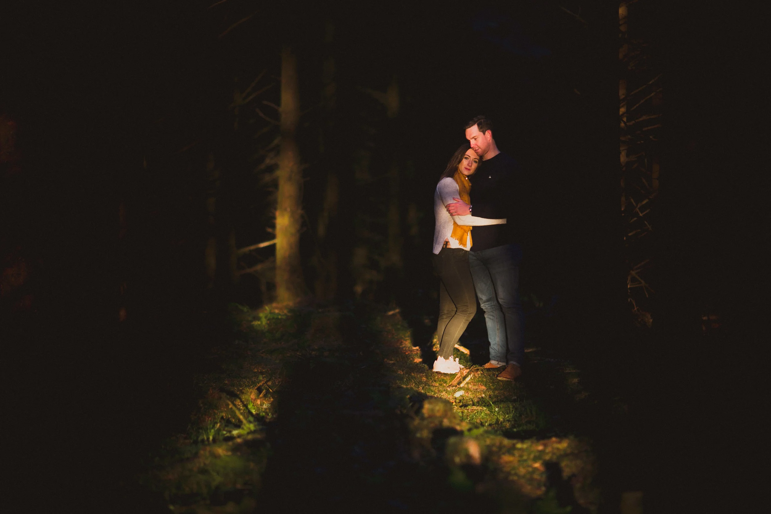 A couple embraces in a dark forest illuminated by a light source on the ground.