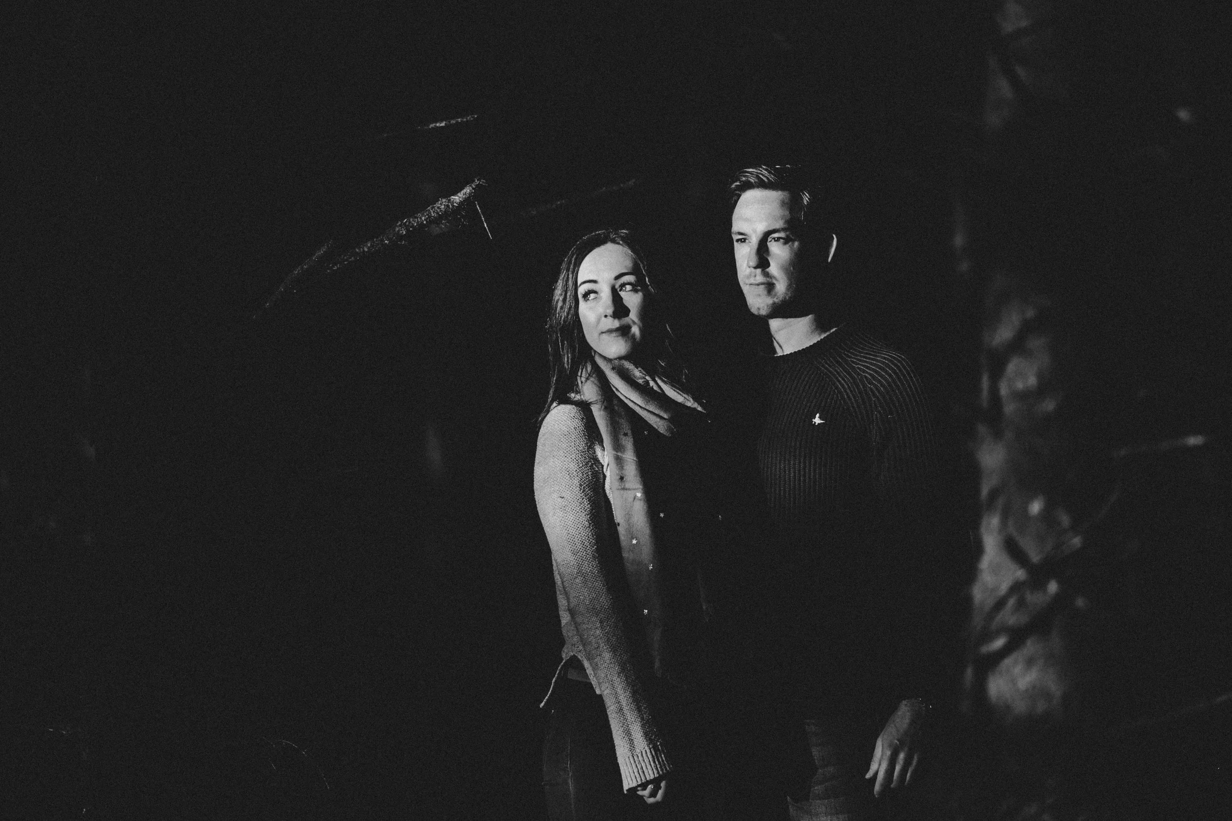 A black and white photo of a man and woman standing outdoors at night with trees in the background.