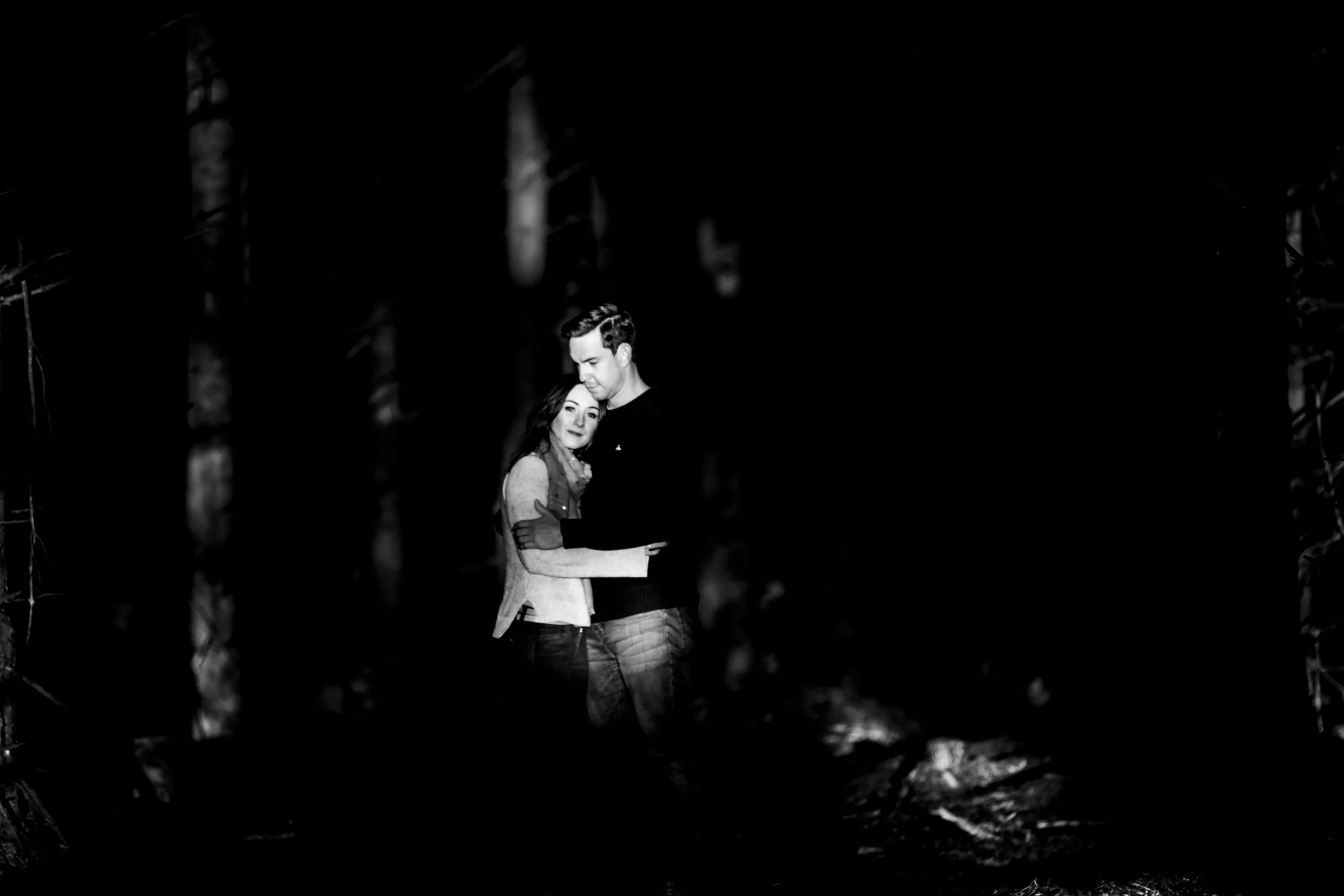 A black and white photo of a couple embracing in a dark outdoor setting, possibly at night, with the woman resting her head on the man's chest. Couples Session
