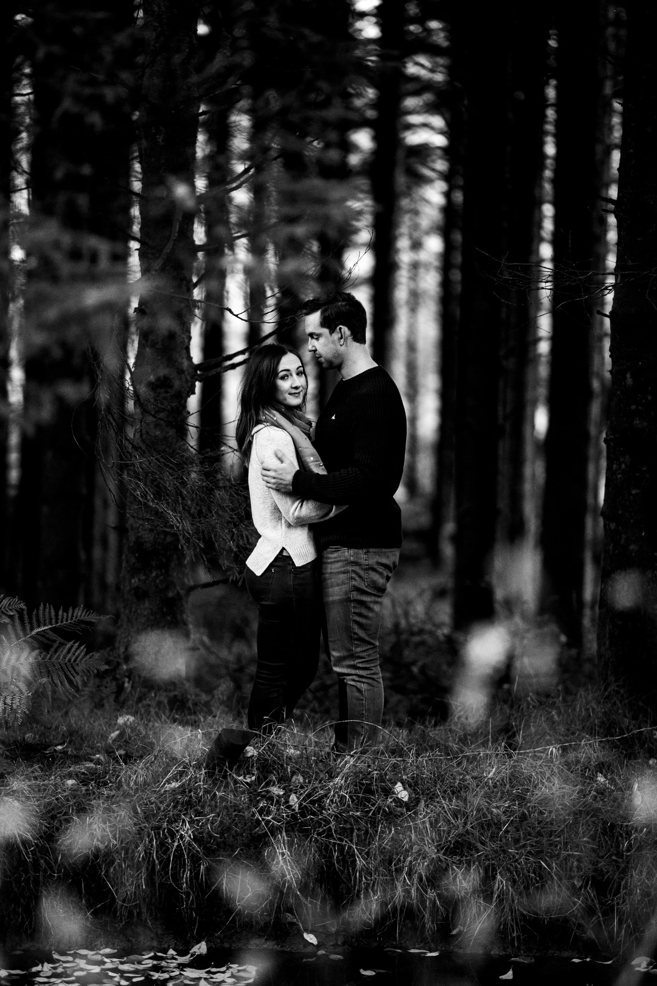 A black and white photo of a couple embracing in a forest, standing on grass near a small body of water, with trees in the background.