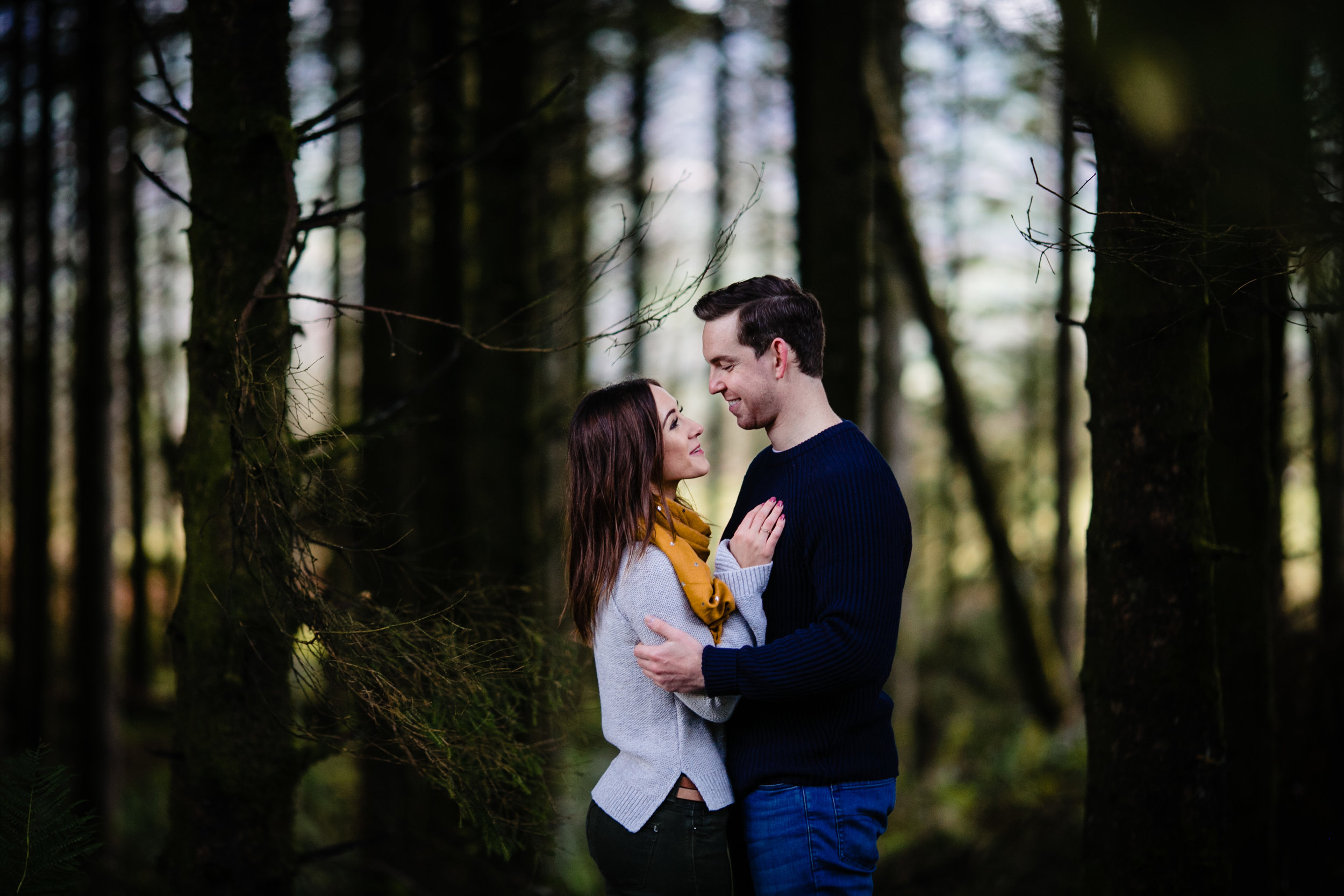 A couple standing close together in a forest, smiling and looking into each other's eyes.