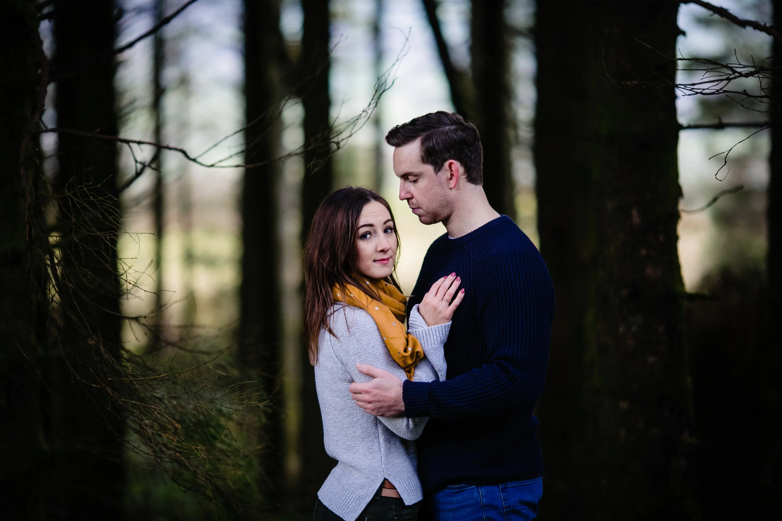 A young couple embracing in a forest with tall trees and blurred background.