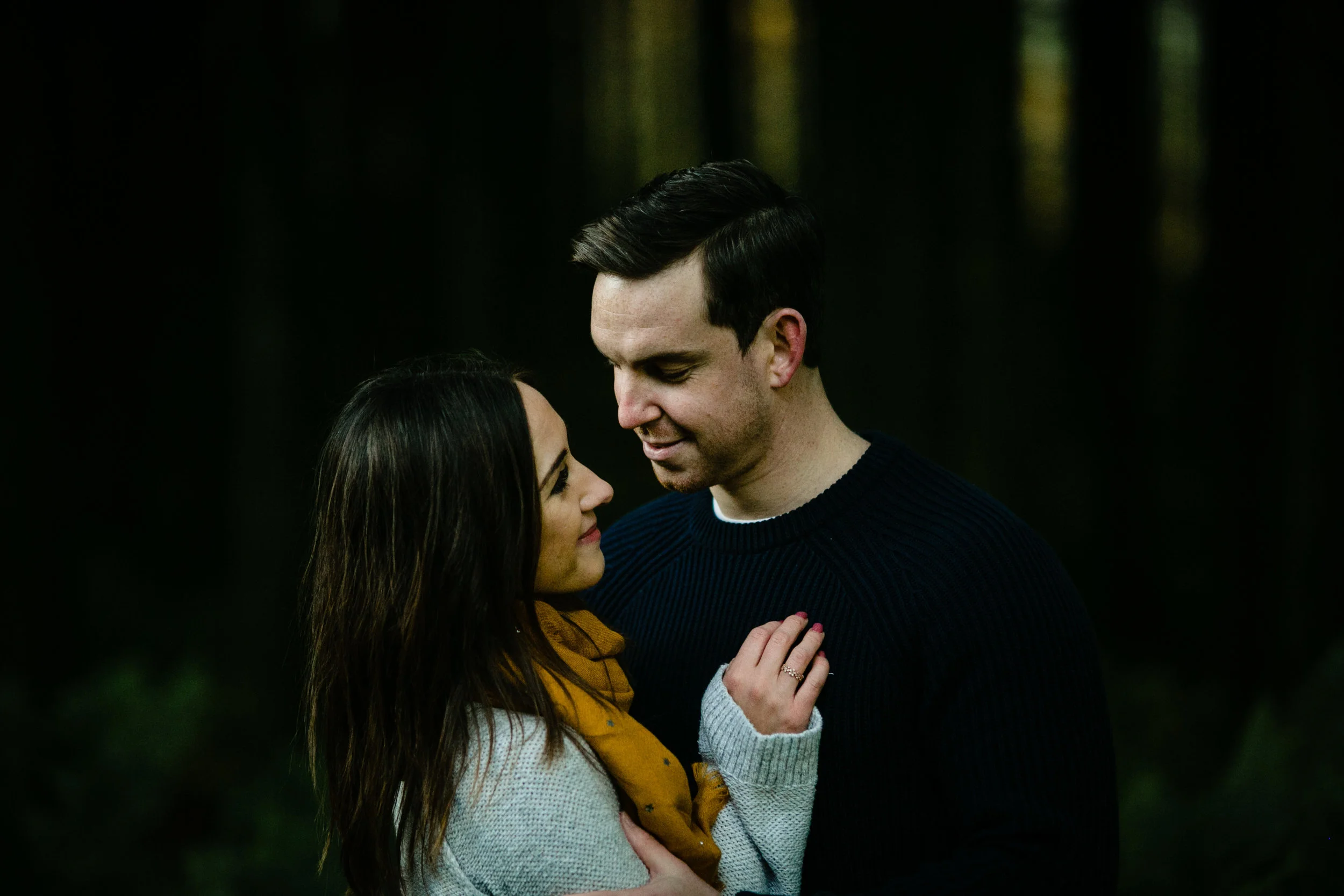 A man and a woman standing close together outdoors at night, gazing at each other affectionately, with a blurred dark forest background.