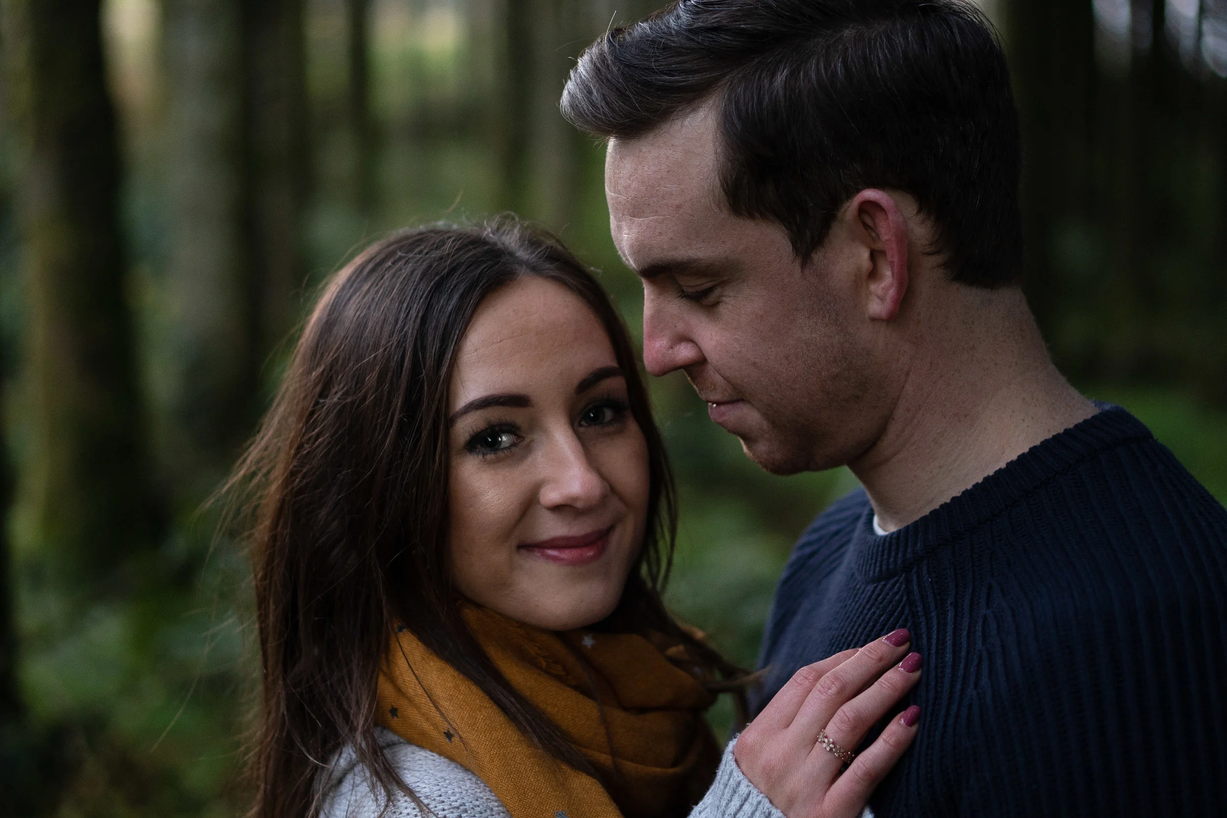 A woman and a man in a forest, close together with the woman smiling and touching the man's chest.