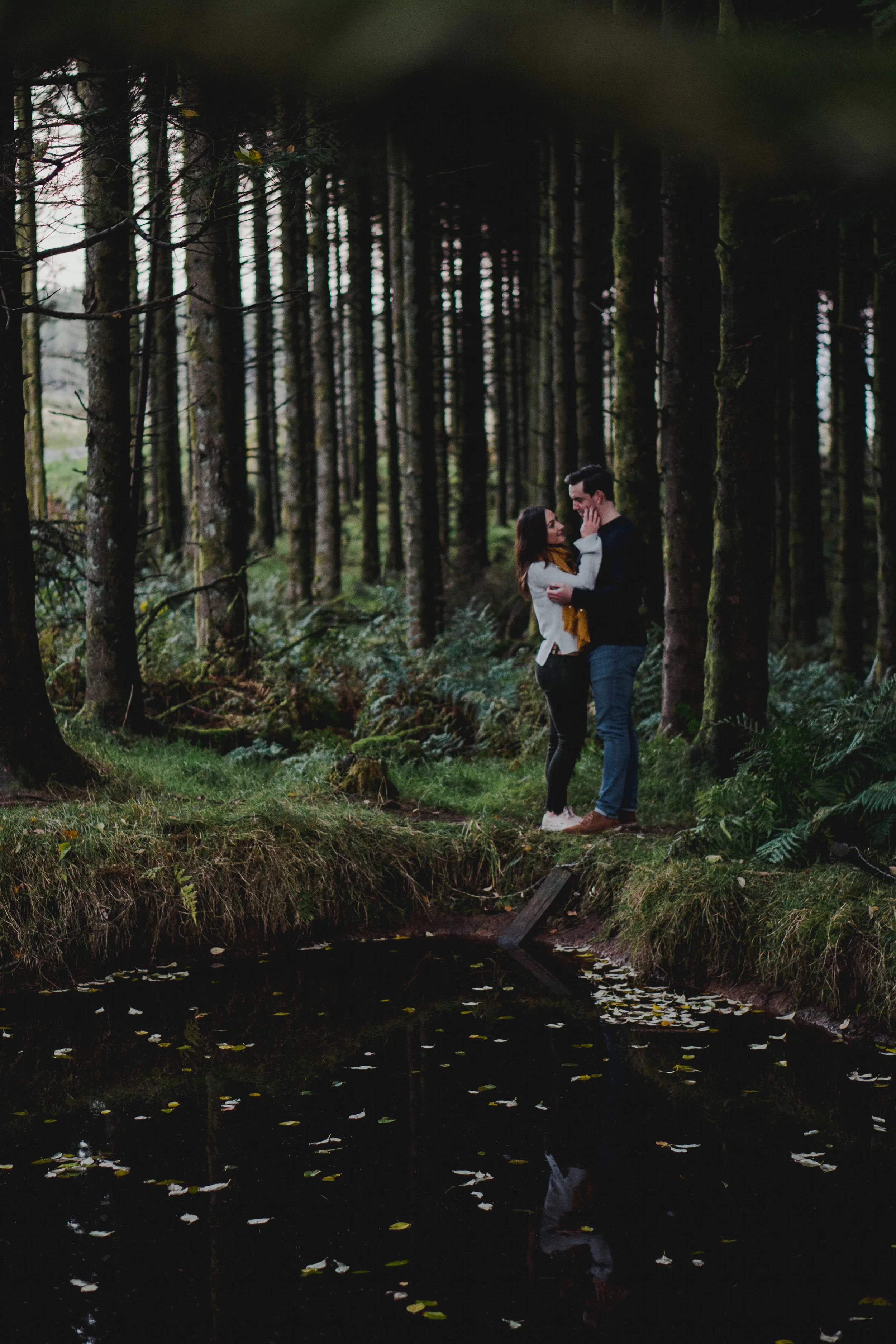 A couple stands close together on a grassy forest path, touching faces, surrounded by tall trees, with a pond reflecting the scene and fallen leaves floating on the water.