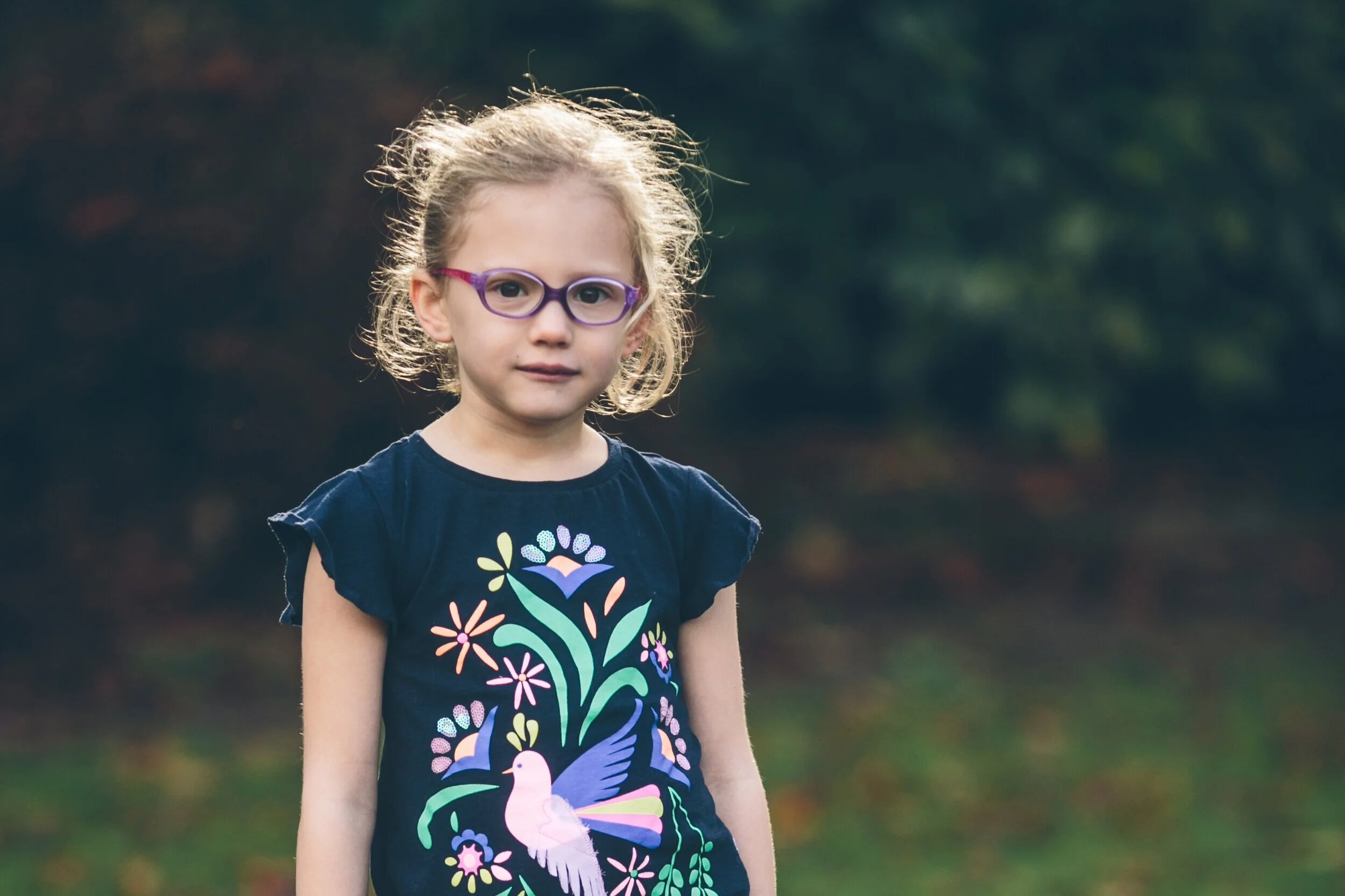 A young girl with light curly hair and purple glasses standing outdoors with a blurred natural background.