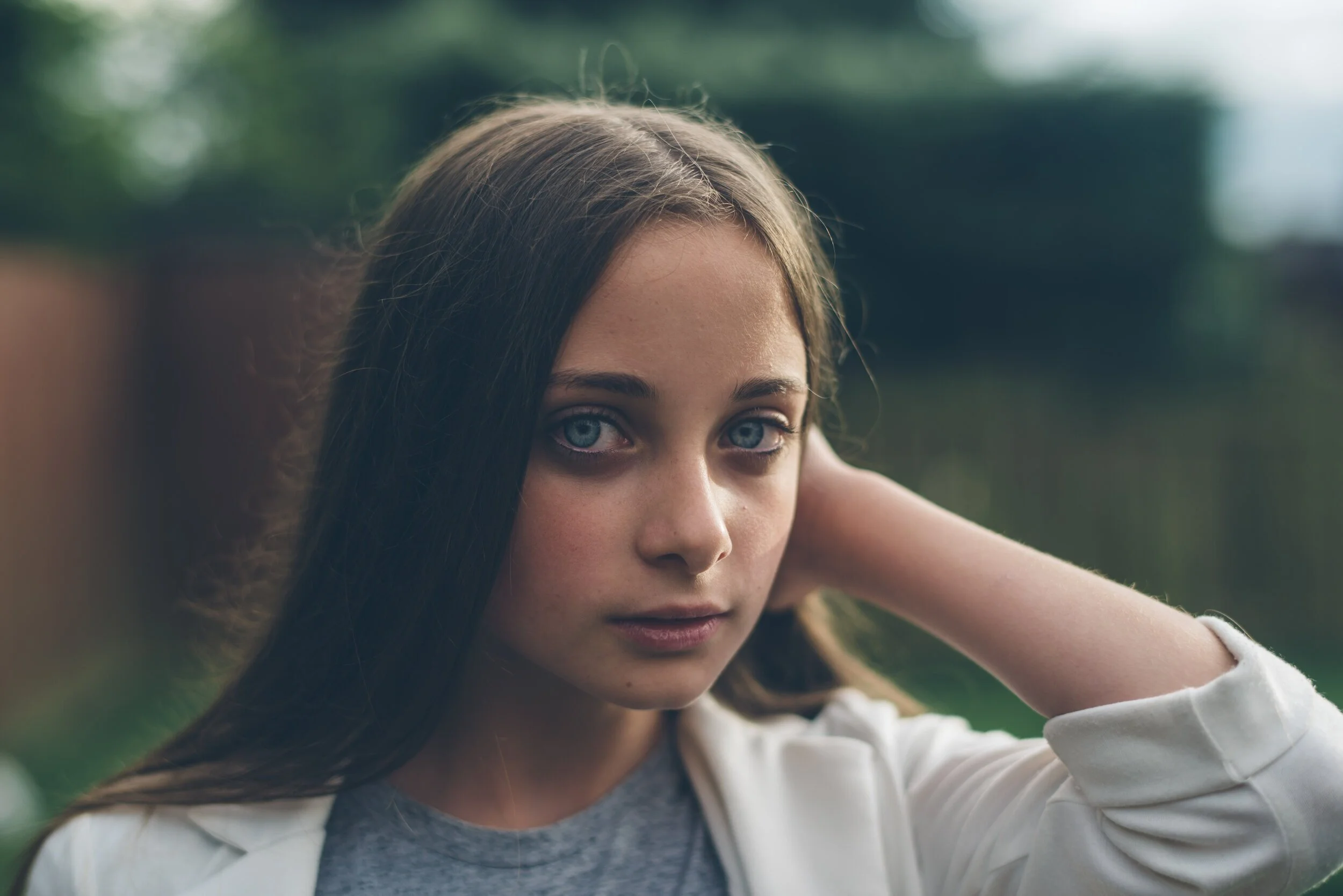 A young woman with blue eyes and long brown hair outdoors, wearing a white jacket and gray shirt, with her hand resting on her neck, looking into the camera.