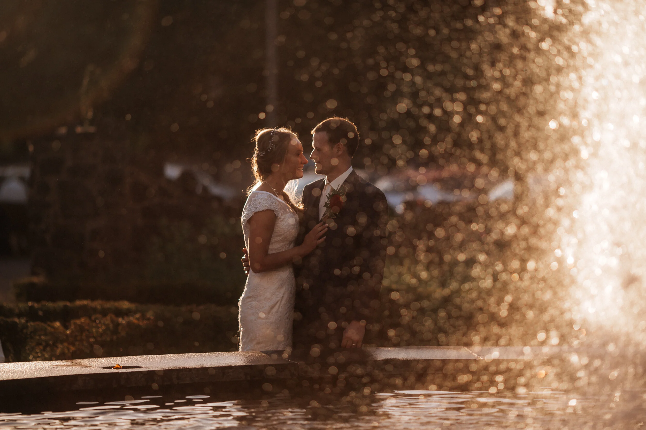 A bride and groom standing close together near a fountain during sunset, with water droplets sparkling around them.