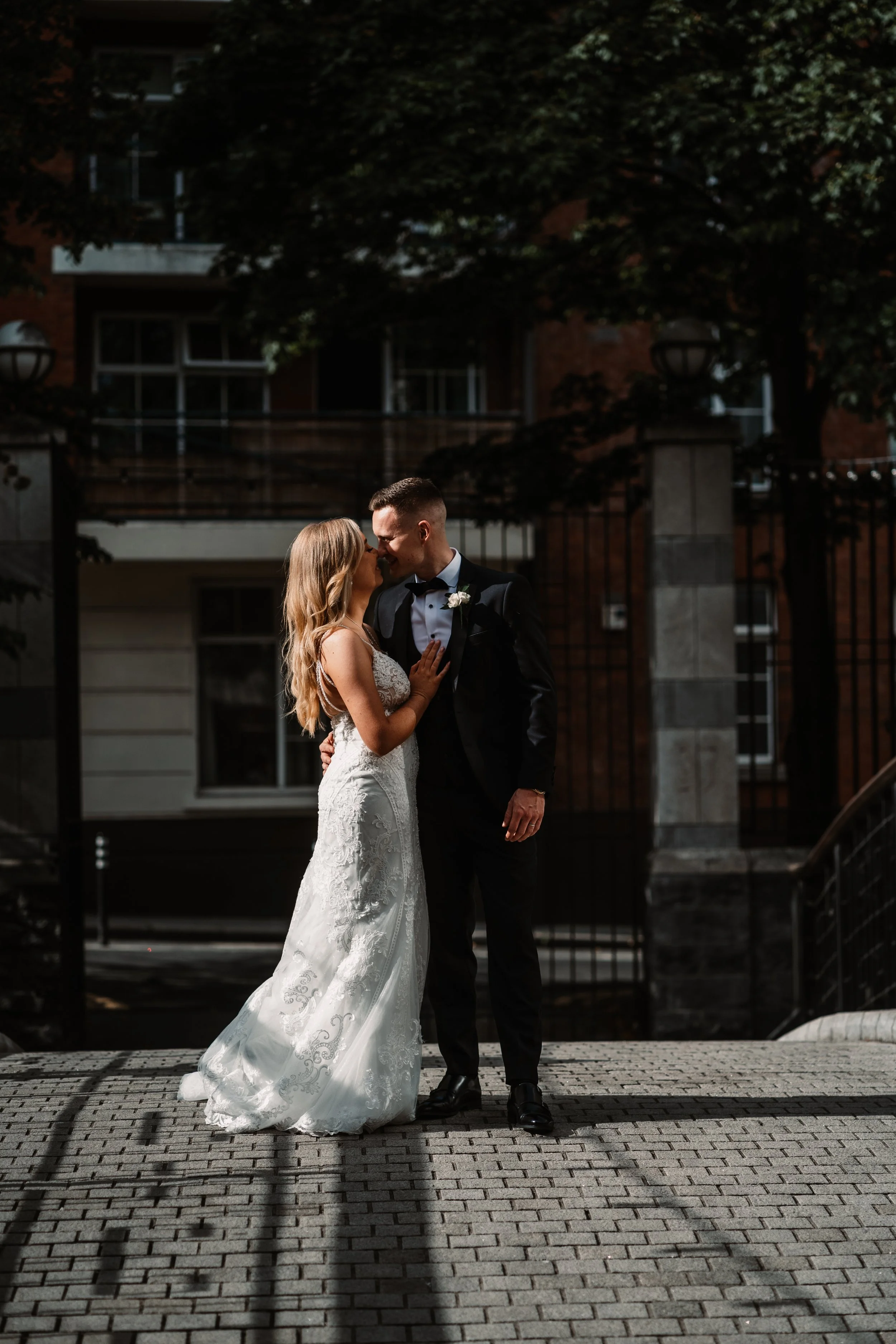 A bride and groom sharing a romantic moment outside, dressed in wedding attire, with the bride in a white lace gown and the groom in a black tuxedo, surrounded by trees and urban buildings. Dublin City Castle