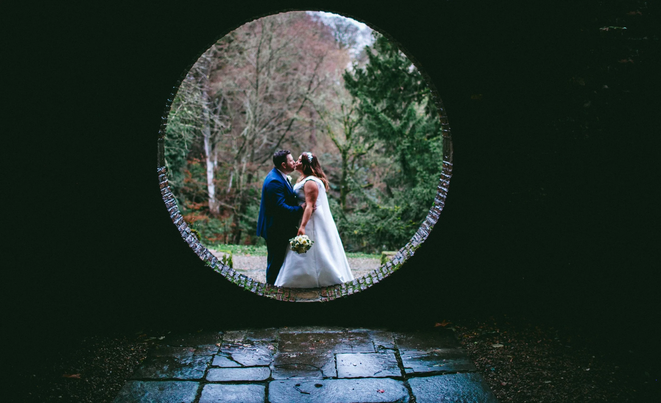 A wedding couple kissing outdoors, viewed through a circular opening in a dark structure, with trees and foliage in the background. Drenagh Estate