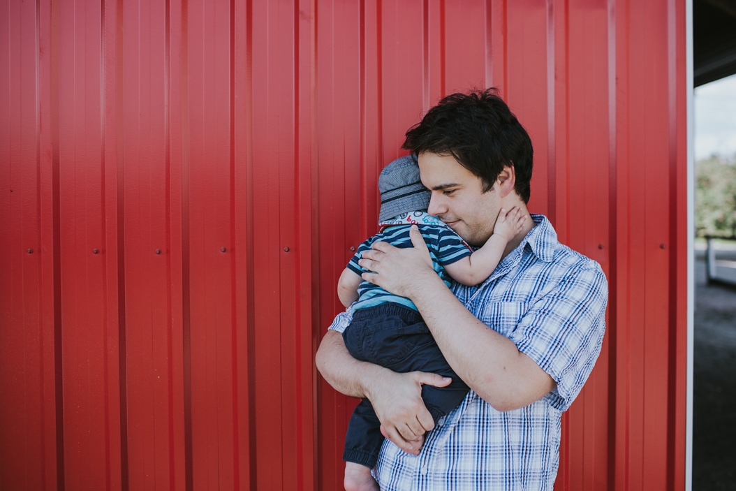  dad holding baby in front of a red wall at apple orchard in ontario canada 