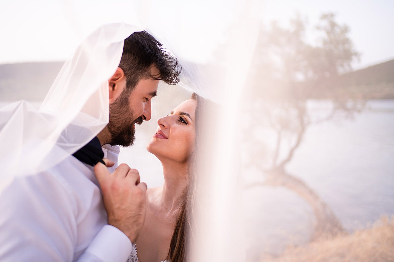  As the sun sets over the picturesque beach, the bride and groom stand beneath a delicate veil, their love for one another evident in their glowing smiles. Surrounded by towering olive trees, they share a moment of peaceful solitude, basking in the b
