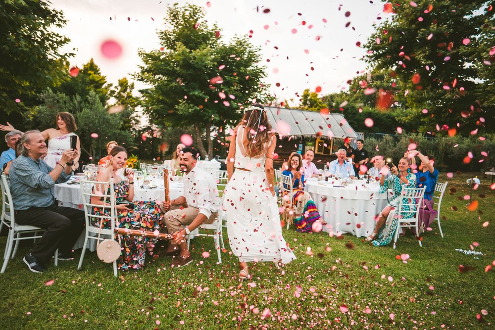  As the sun sets on the horizon, casting a warm glow over the landscape, a beautiful civil marriage ceremony takes place. The bride stands with her back to the camera, her white dress contrasting beautifully with the vibrant confetti that surrounds h