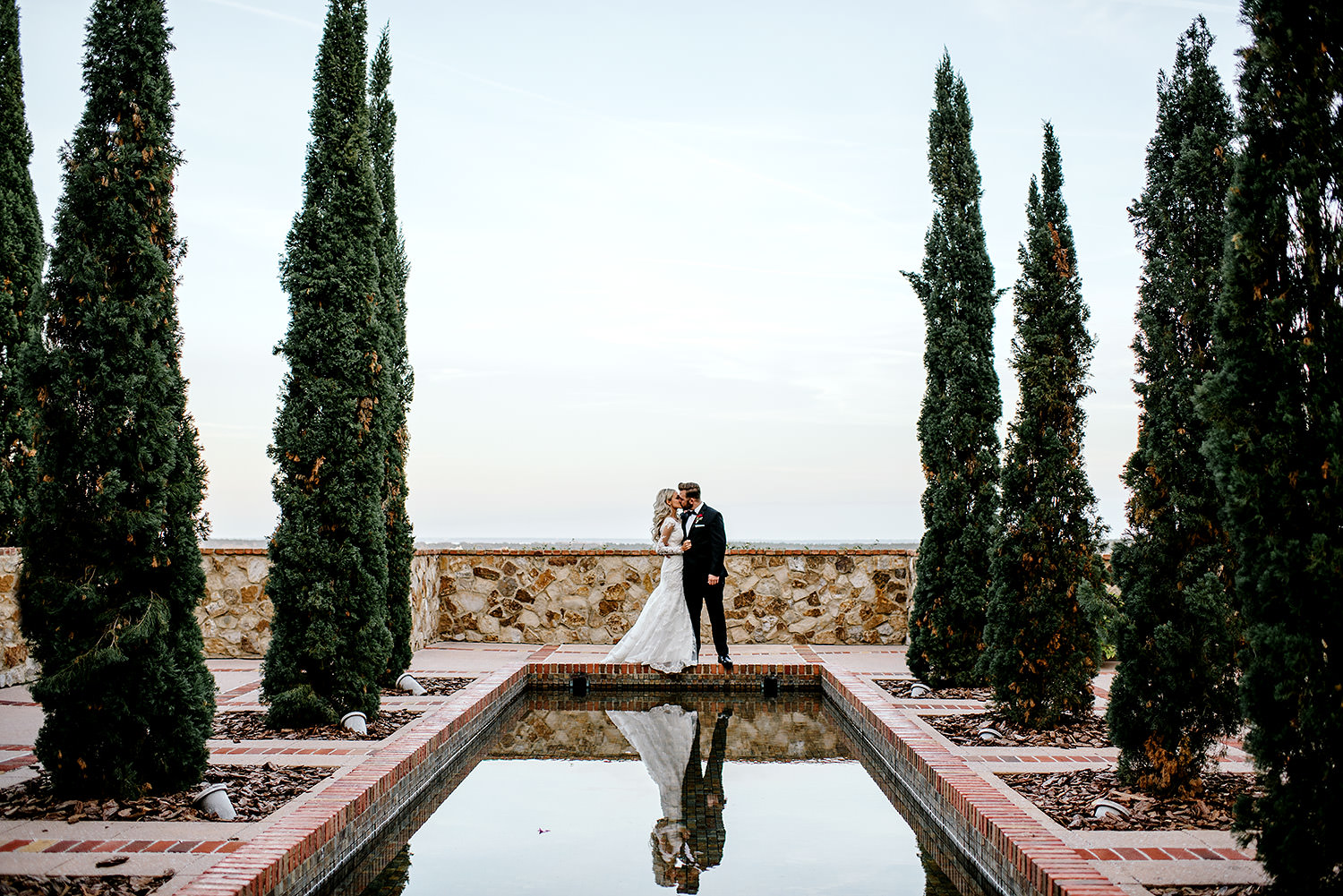 Bride and groom kissing in front of beautiful pool outlined with tall spartan juniper trees at Bella Collina Florida.