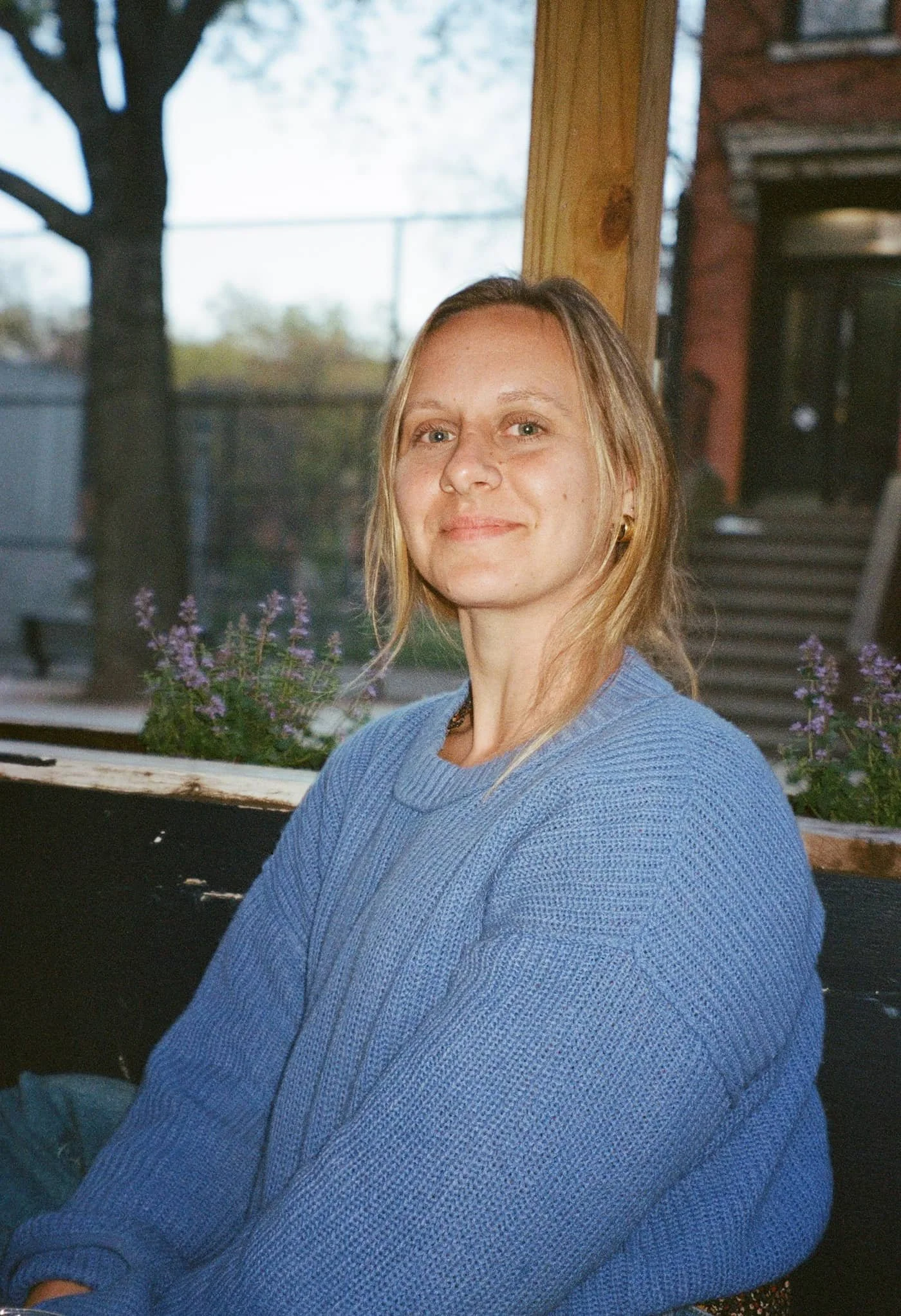 Image of Amelia Broussard, A woman with long blonde hair and green eyes wearing a blue sweater, sitting near a window with a view of trees and a fence outside, with purple flowers in the foreground.