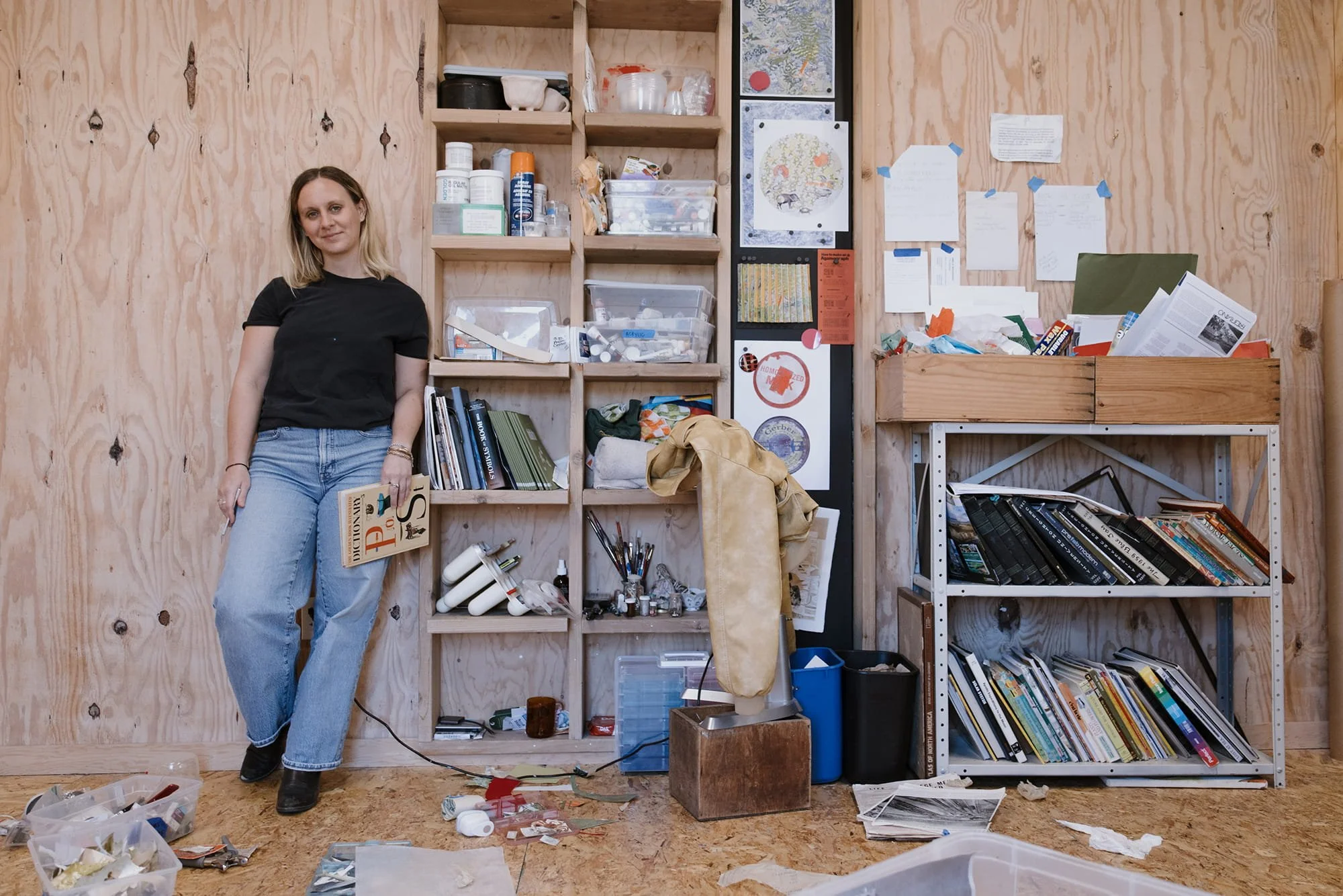 woman with shoulder-length blonde hair dressed in a black t-shirt and light blue jeans stands in a cluttered workspace with unfinished wooden walls and shelves filled with various supplies, books, and materials. The floor and nearby surfaces are scattered with papers, tools, and art supplies.