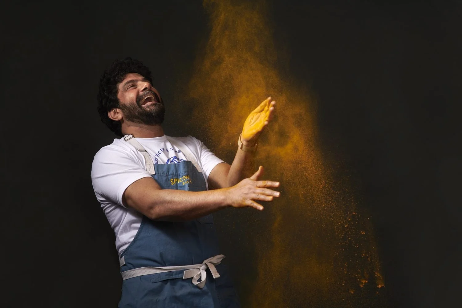 A chef in a denim apron throws his head back laughing as a cloud of orange turmeric hangs in the air against a black backdrop.