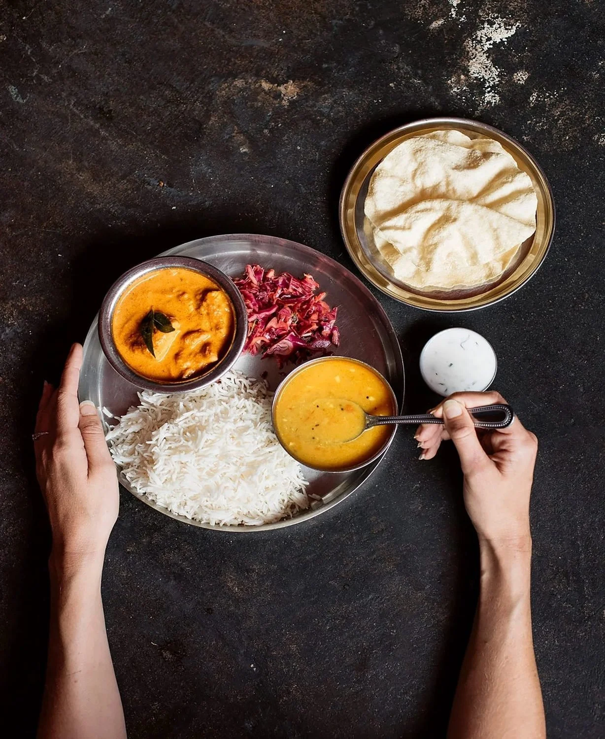 Overhead of a steel thali with rice, dal, curry and red cabbage on dark stone, hands holding the plate and a spoon.