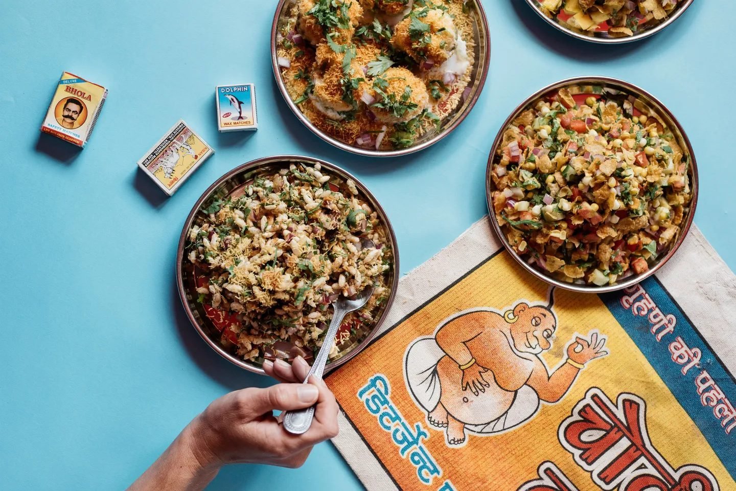 Overhead of three plates of chaat on a pale blue surface with vintage matchboxes and a printed cloth, a hand scooping with a spoon.