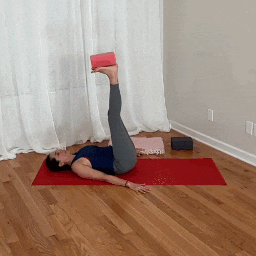 Supta padangusthasana variation balancing a yoga block on the foot while lowering the opposite leg.