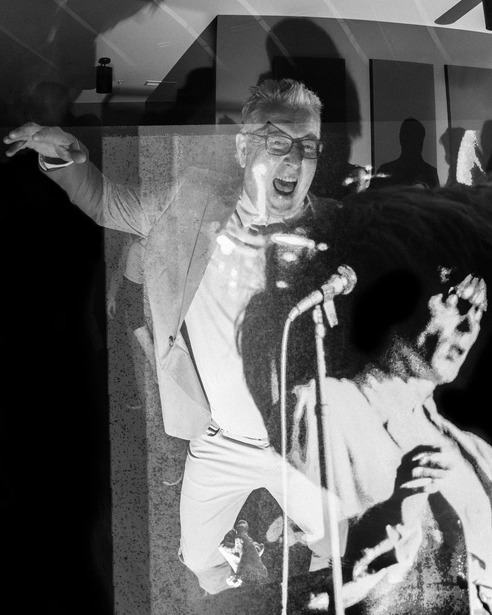Black and white photo showing a man with glasses and gray hair passionately speaking or shouting into a microphone, surrounded by an audience and reflections in the background.