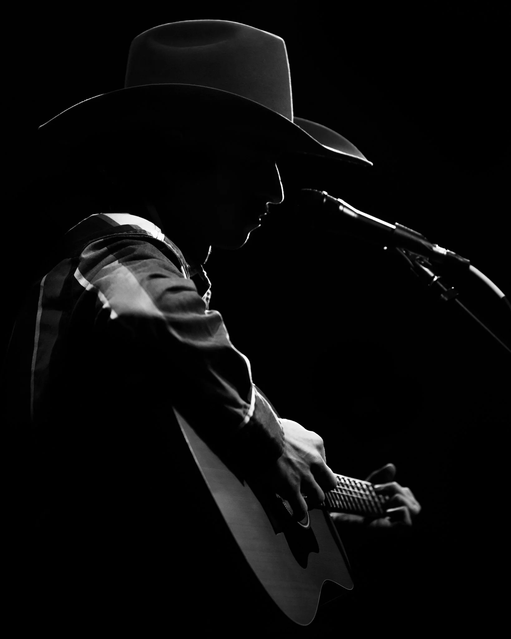 Silhouette of a person wearing a cowboy hat playing an acoustic guitar against a black background.
