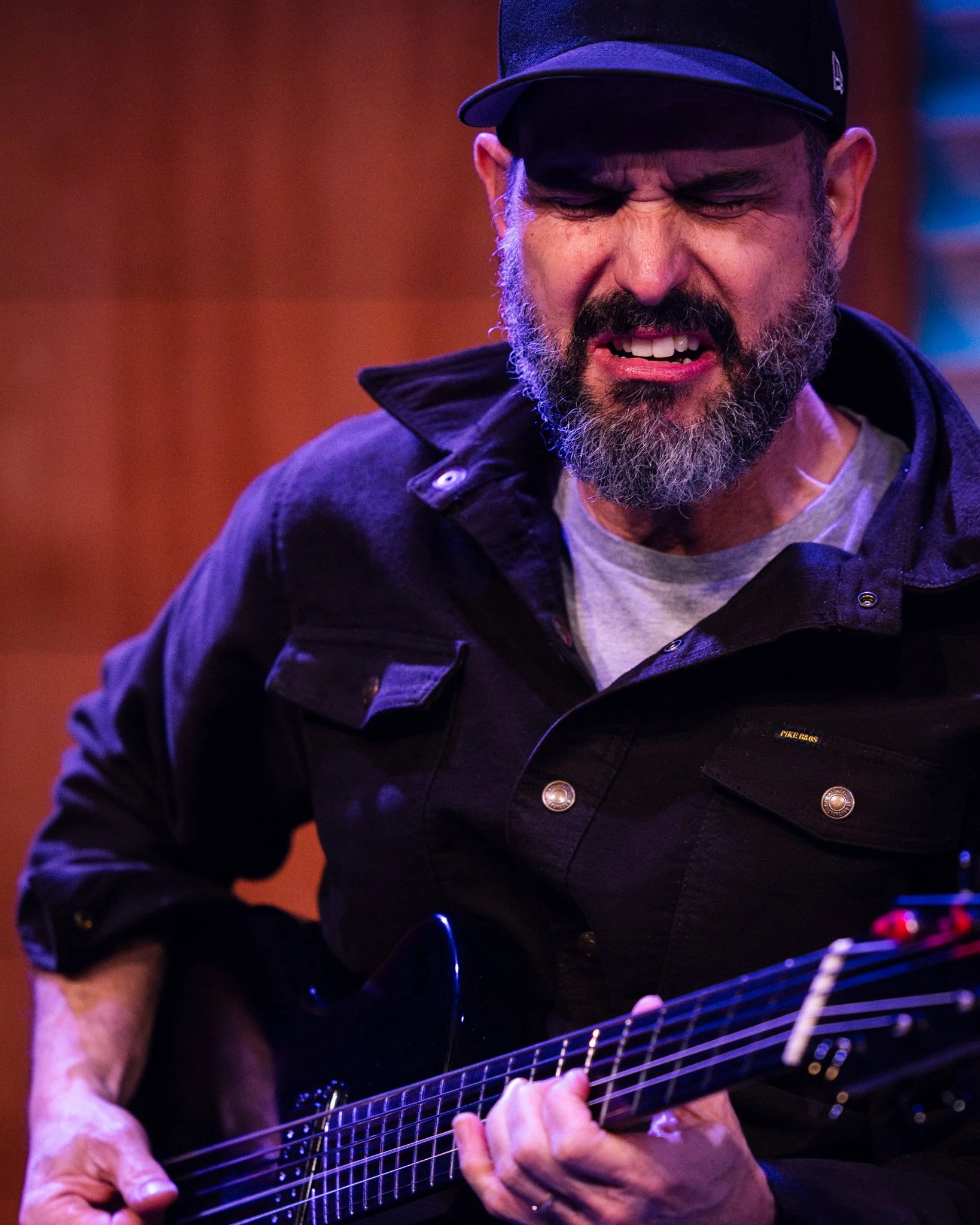 A man with a beard and wearing a black cap and jacket playing an electric guitar, with an expressive facial expression.