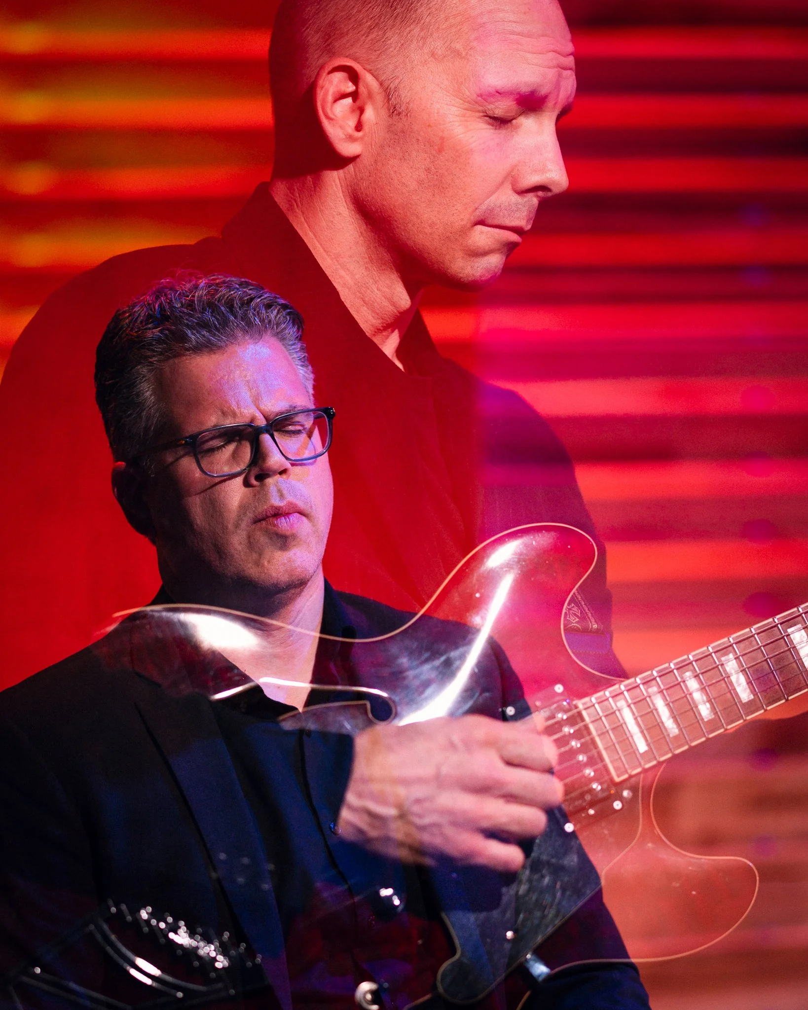 Two men, one playing an electric guitar, with a red background and colorful streaks of light.