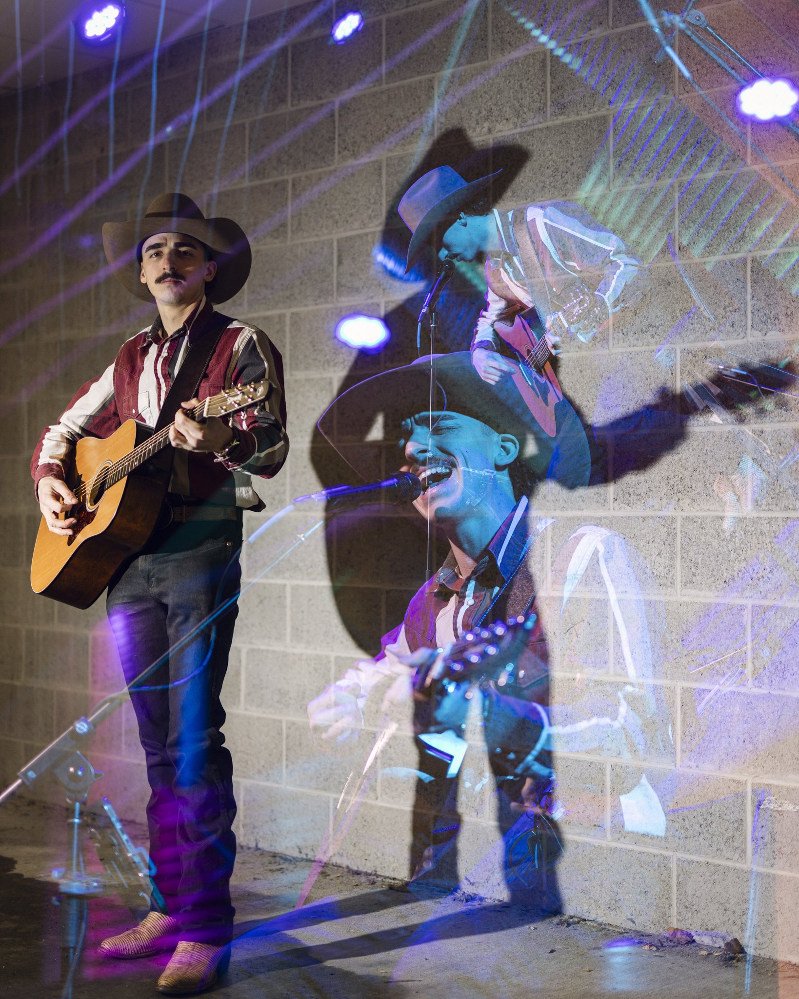 A man in a cowboy hat and striped shirt plays an acoustic guitar, while another man with a cowboy hat and striped shirt sings and plays an acoustic guitar. They perform against a brick wall with colorful light projections.