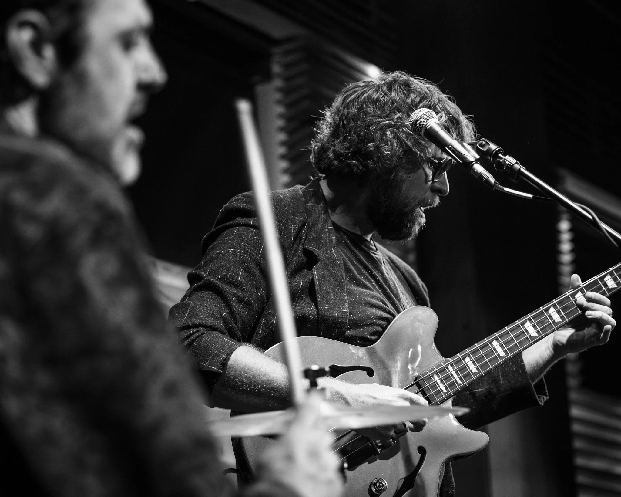 Two musicians perform on stage; one playing drums, and the other playing an electric guitar, singing into a microphone, in black and white.